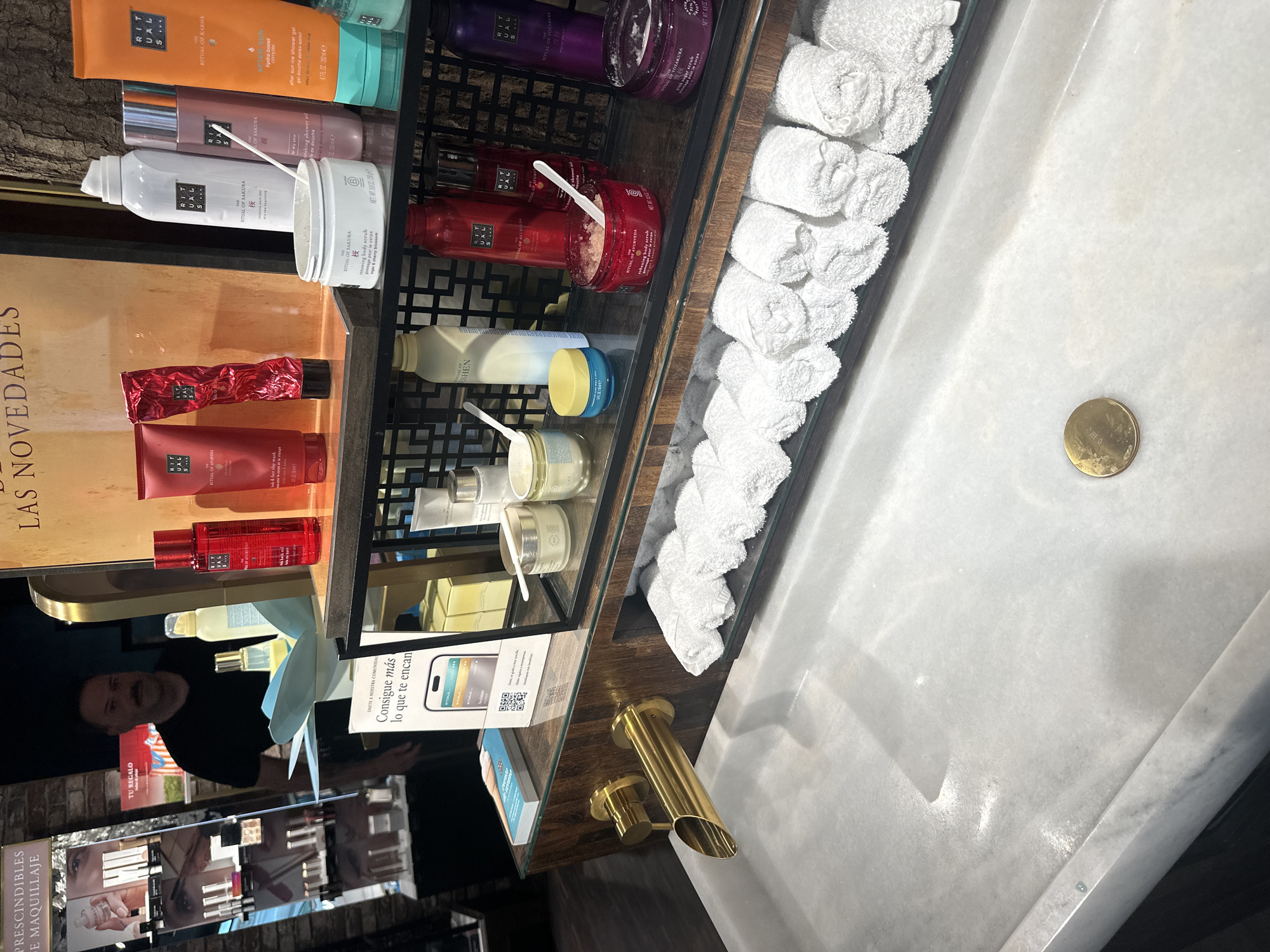 This image depicts a product display, likely within a store setting. The display features a multi-tiered black shelving unit holding a variety of personal care products from the brand Rituals. These include shower gels, body scrubs, lotions, and other cosmetic items. Each shelf is neatly arranged, with some products featuring small white spoons for sampling.
Adjacent to the shelving unit is a white marble sink. Rolled white towels are positioned along the edge of the shelf, next to the sink, suggesting a spa or beauty store environment. The sink has a gold-colored faucet and a round gold drain cover.
A reflection in the glass surfaces of the display shows a person with a mustache wearing a black shirt, as well as various marketing materials and product advertisements. The language on the displays is a mix of English and Spanish, indicating a possible location in a region with a multilingual population. The environment appears to be a modern retail space with attention to cleanliness and aesthetics.