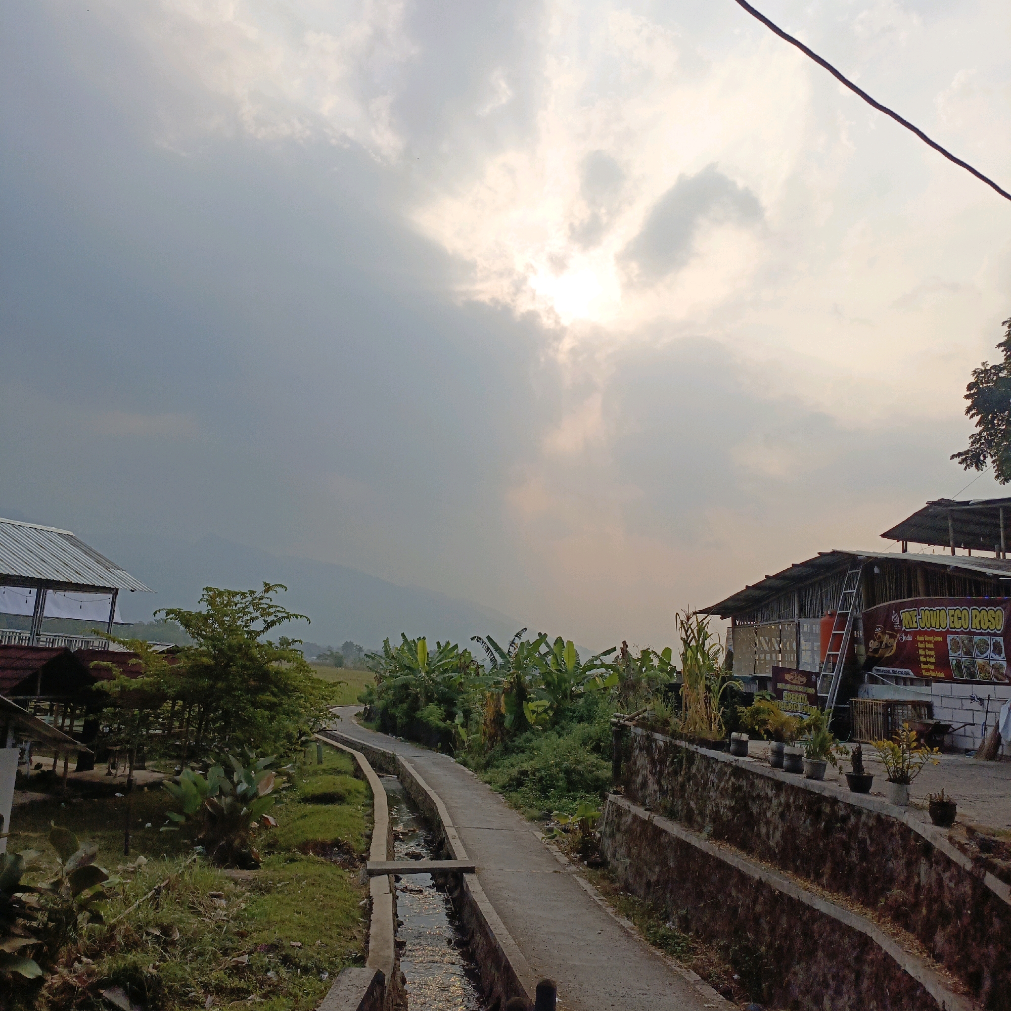 This image depicts a rural or semi-rural scene in Salatiga, Indonesia, likely near a roadside eatery or rest stop. The sky is overcast with the sun partially obscured by clouds, creating a diffused light. In the foreground, a concrete path runs alongside a narrow canal or drainage ditch filled with water. The path is bordered on the right by a textured stone wall supporting a raised area with several potted plants.
To the right of the path, there are buildings with corrugated metal roofs, characteristic of roadside establishments or simple homes. A banner with Indonesian text and images of food is visible on one of the buildings. A ladder leans against one of the structures. Various types of vegetation are present, including banana trees and other leafy plants, along with some dry stalks of corn.
On the left side of the image, there are more structures with corrugated roofs, including what appears to be an open-sided shelter with white curtains. More lush greenery and trees surround these buildings. In the distance, hills or mountains are faintly visible through the haze. The overall impression is a functional, possibly commercial, area integrated with the natural landscape.
