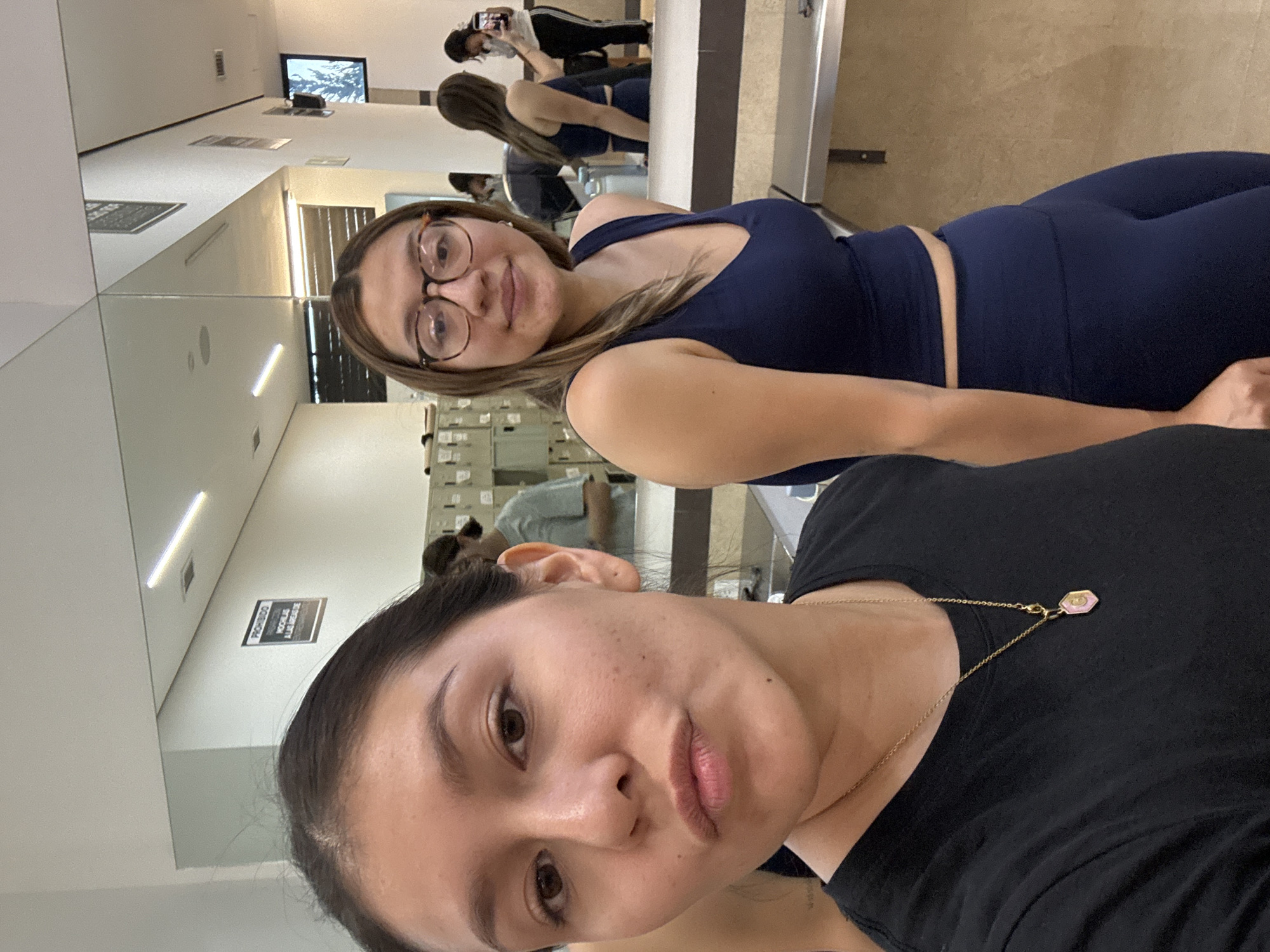Two women pose for a selfie. The woman in the foreground is wearing a black shirt and gold necklace, while the woman in the background is wearing a blue athletic outfit and glasses. They are in a mirrored room that appears to be a gym or studio. The location is identified as San Andrés Cholula, Mexico based on the reflections in the mirrors.