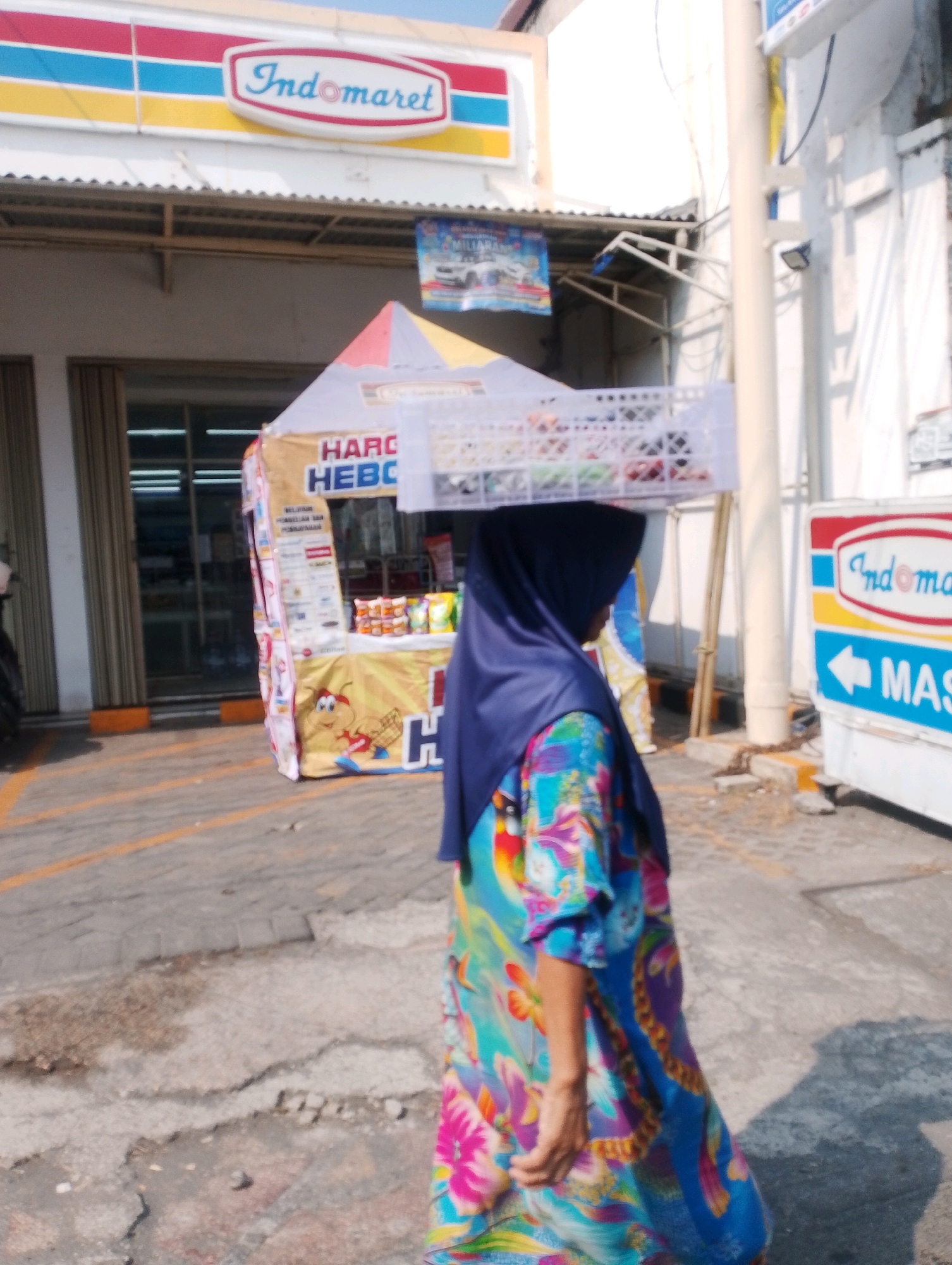 The image shows a woman wearing a blue hijab and a colorful patterned dress walking in front of an Indomaret convenience store in Probolinggo, Indonesia. She is carrying a white plastic crate filled with items on top of her head. The Indomaret store has its characteristic red, blue, and yellow logo visible. To the right of the store, there is a sign with an Indomaret logo and an arrow pointing to the left. A small, tent-like kiosk with advertisements is set up in front of the store. The ground is paved with concrete and asphalt, with some cracks and debris visible. The lighting suggests it is daytime, with sunlight casting shadows.