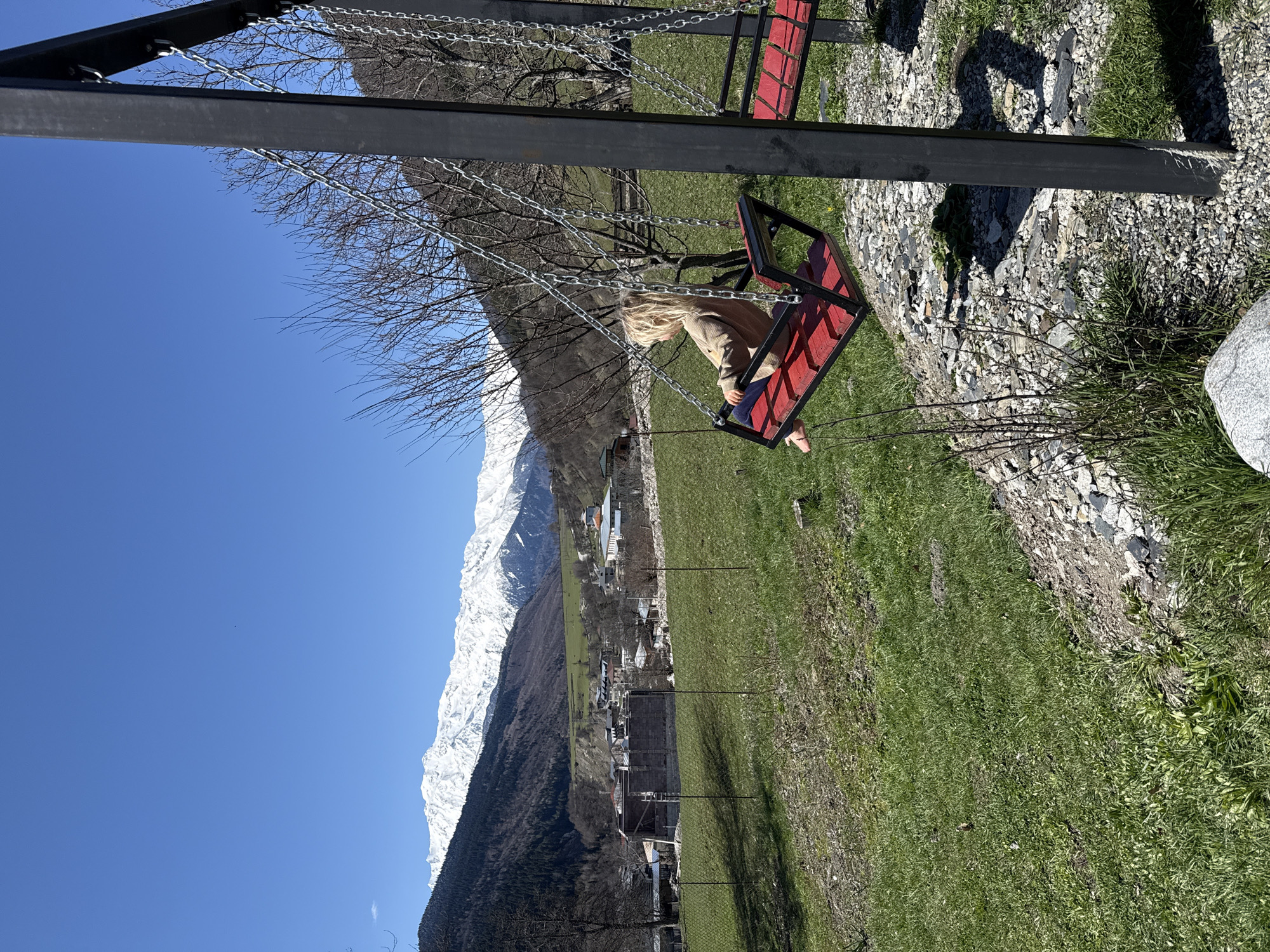A serene outdoor scene captured on a bright, clear day in Mestia, Georgia, features a child on a swing set against a backdrop of snow-capped mountains. The image is oriented sideways, but should be viewed as if the horizon is horizontal.
In the foreground, a vibrant green grassy area is mixed with patches of light-colored gravel and rocks. A dark metal swing set frame supports a red-seated swing. A child with long blonde hair, wearing a light brown or beige jacket/cardigan and dark pants, is seated on the swing, facing away from the viewer and towards the distant mountains. Their feet, clad in light-colored shoes, are visible.
Beyond the immediate foreground, several bare trees suggest a season of late winter or early spring. A small village is nestled in the midground, with various buildings visible among the trees at the base of the mountain.
The background is dominated by a majestic range of towering, snow-capped mountains that stretch across the horizon under a perfectly clear, bright blue sky. A distinct treeline marks the transition from dark forested slopes to the white, snowy peaks. The bright sunlight indicates it is daytime, casting clear shadows on the ground. The scene conveys a sense of peaceful activity amidst a grand natural landscape. No text is visible in the image.