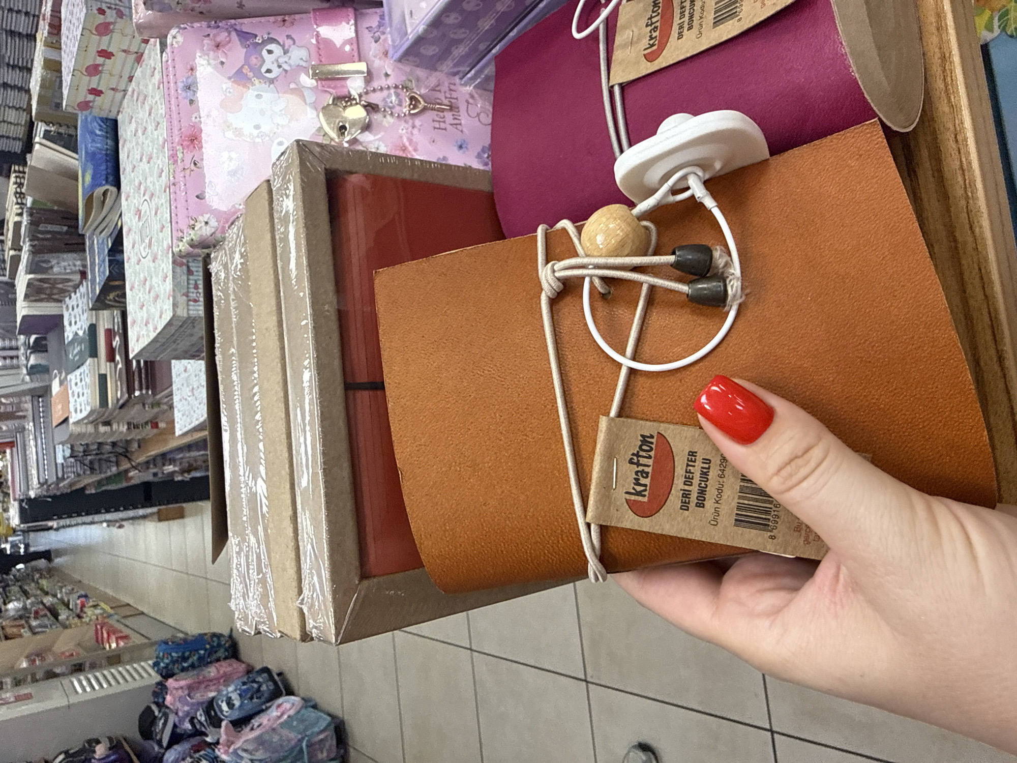 A woman's hand, with bright red nail polish, holds an open brown leather-bound notebook. A string with wooden beads and black stoppers wraps around the notebook, securing it. A Krafton brand tag is attached to the string, with Turkish text indicating "Leather Notebook with Beads" and a product code. The notebook is positioned in front of a purple notebook or folder. Behind these items, a cardboard box wrapped in plastic contains more notebooks or similar items. In the background, shelves are stocked with various stationery items, including backpacks, boxes, and books, suggesting a retail environment like a stationery store or gift shop. The floor tiles are visible. The scene appears to be indoors, likely in Kadıköy, Turkey, given the presence of Turkish text on the product tag.