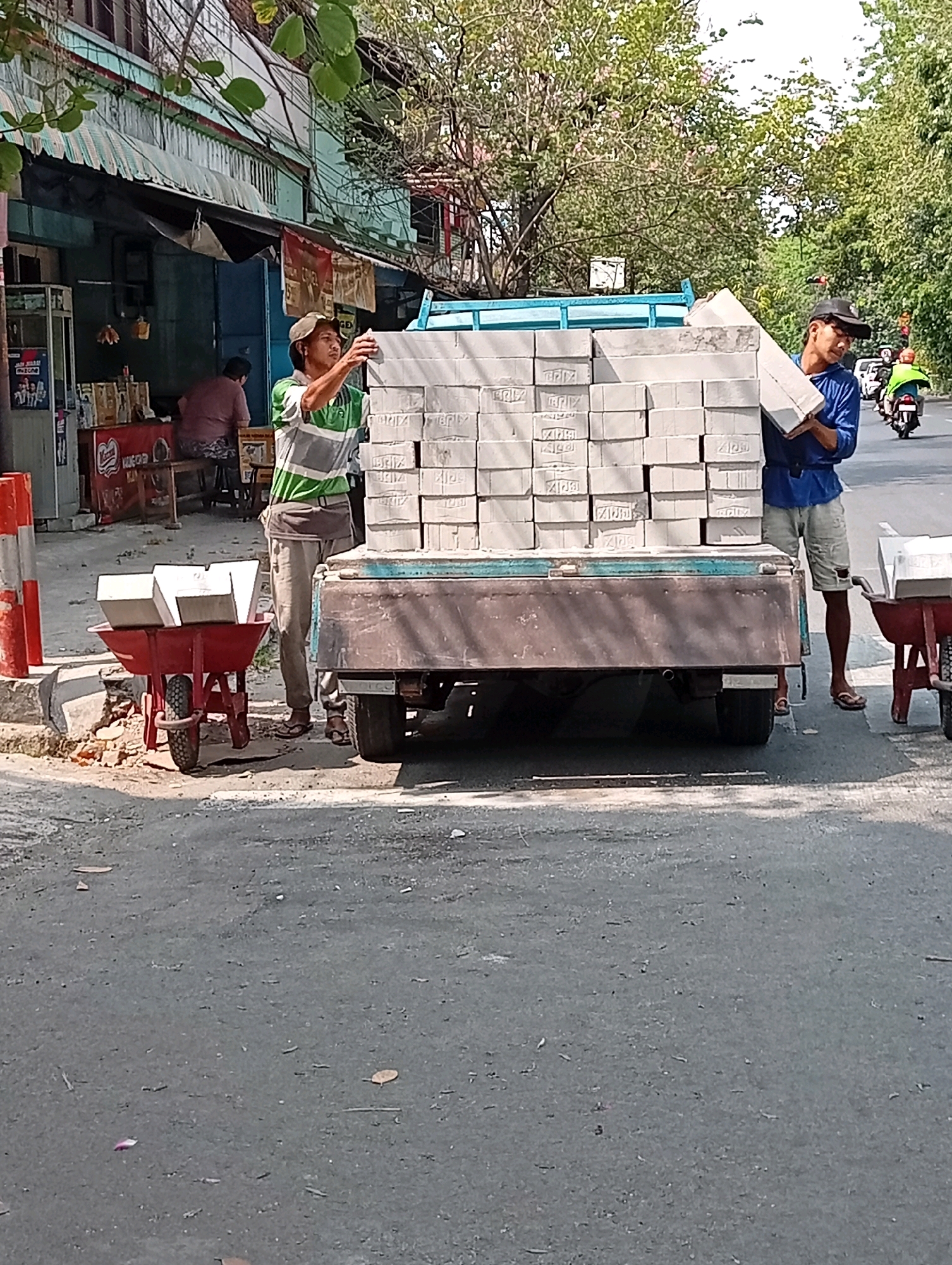 The image depicts two men loading bricks onto a flatbed truck on a city street in Surabaya, Indonesia. The scene appears to be in front of a storefront with a light blue facade and a corrugated metal awning. The street is paved and shows signs of wear.
One man, wearing a green and grey striped shirt, a brown belt, and a light-colored cap, is standing on the left side of the truck. He is reaching for a brick from a stack on the truck bed. In front of him, on the sidewalk, is a red wheelbarrow containing two bricks.
The second man, wearing a blue long-sleeved shirt, light-colored shorts, and a dark baseball cap, is on the right side of the truck. He is holding a brick and appears to be placing it onto the stack. A second red wheelbarrow with bricks is visible on the right side of the frame.
In the background, there is a person sitting at a table inside the storefront, and further down the street, a motorcycle with a rider wearing a bright green vest is visible, suggesting a busy urban environment. Lush green trees are present along the side of the road and behind the buildings. The lighting suggests it is a sunny day.