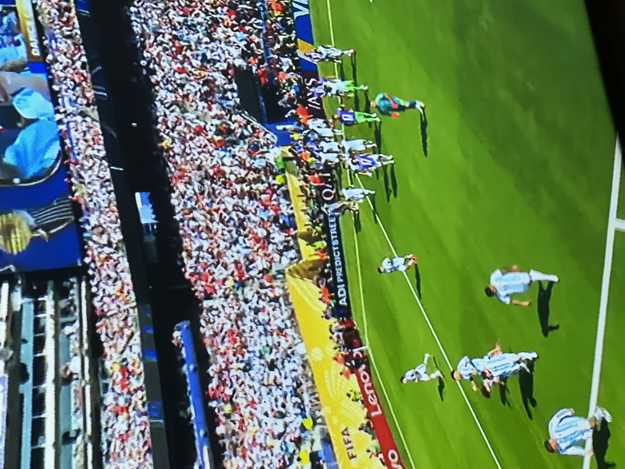 The image depicts a soccer match from a high-angle perspective, showing players on a bright green field. The stadium is packed with a large crowd, with spectators in white and red attire filling the stands. Several advertising banners are visible along the edge of the field, including "FIFA", "ADI PREDICTSTREET", and "Lenovo". Some players are wearing light blue and white striped jerseys, while others are in white or green. One player in the foreground is wearing a green jersey and orange gloves, suggesting they are a goalkeeper. The overall scene is vibrant and energetic, characteristic of a professional sports event. The stadium's architecture suggests a modern design. The image does not contain any explicit geographical markers to pinpoint its exact location within Moscow, Russia.