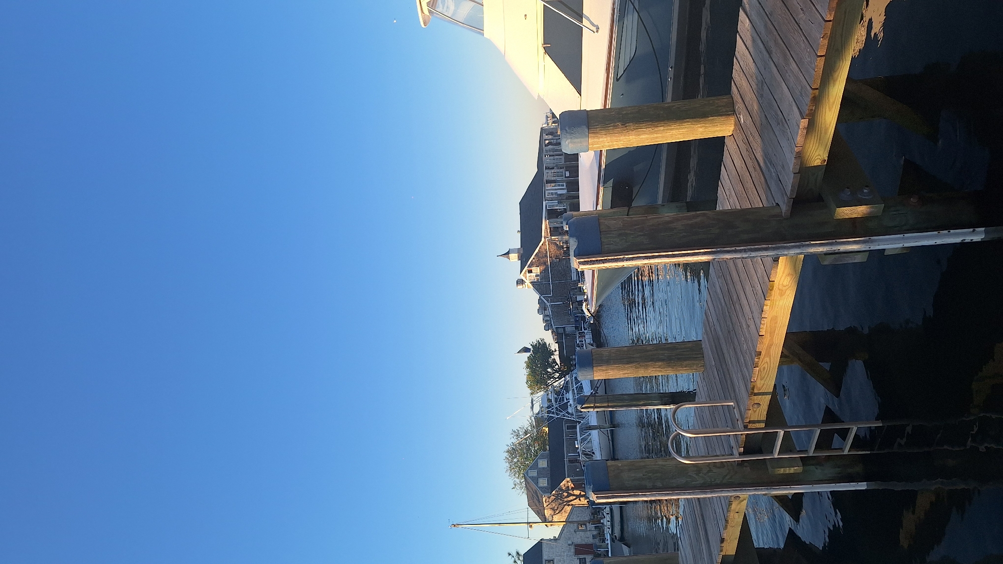 An outdoor vertical shot captures a serene harbor scene on Nantucket, Massachusetts, USA, under a clear, bright blue sky. In the foreground, a wooden dock extends into calm, dark water, with several wooden pilings topped in blue visible. A metal ladder is attached to the dock and submerged in the water. Beyond the dock, several boats are moored in the harbor, including a sailboat with its mast raised and a larger white motor yacht. In the background, a row of charming Nantucket-style buildings with clapboard siding and distinctive steep roofs line the waterfront. The sunlight casts long shadows, indicating it is either early morning or late afternoon. No people are visible in the frame.
