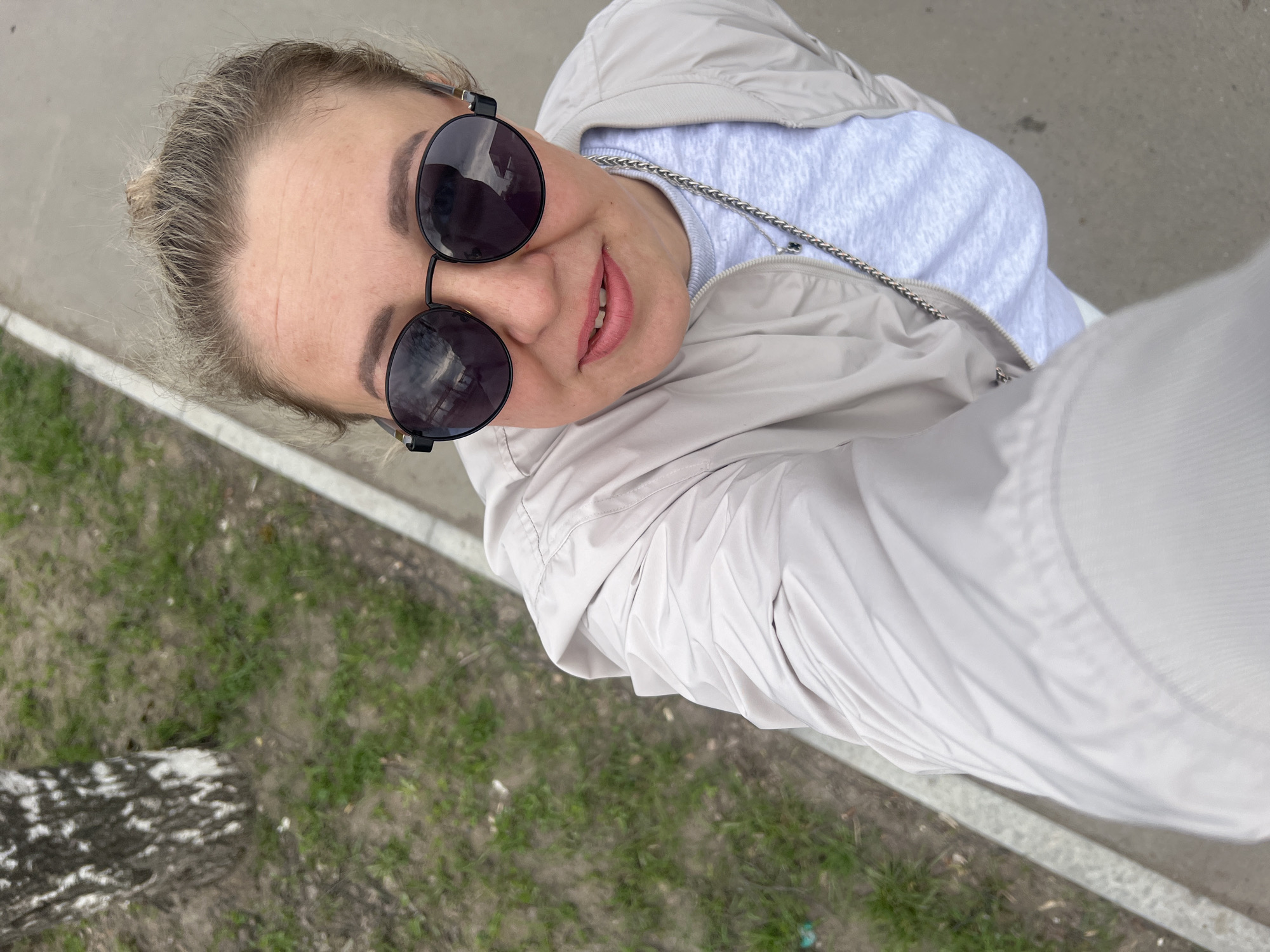 A person, likely female, is captured from a high angle in an outdoor setting. The individual has fair skin, blonde hair styled in a bun, and wears sunglasses with round, dark lenses. She's dressed in a light gray zip-up jacket over a light blue shirt with a chain necklace. The background shows a sidewalk, grass, and a partially visible tree trunk with black and white bark. The setting is urban and likely a park.
It is not possible to verify the location in the city of Chaikovsky, Russia, without further information.