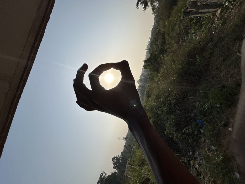 A silhouetted hand makes the “OK” sign, framing the sun. The bright sun shines through the hole created by the fingers. The arm extends upwards from the lower right. In the background, lush green vegetation covers a hillside under a bright, hazy sky. A building edge is visible in the upper left. The overall composition is taken in the middle of the day with the sun shining above the foliage.