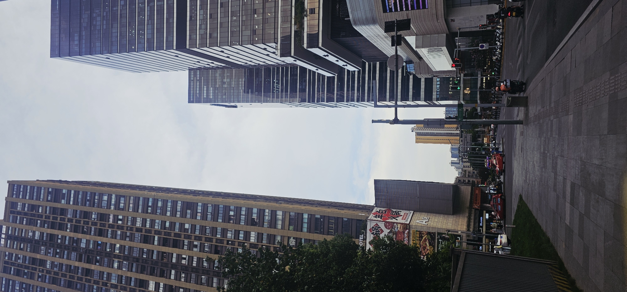The image shows a street scene in Jiulongpo District, China. The perspective is from a sidewalk looking down a street lined with tall buildings. On the left, a very tall, narrow building with many windows dominates the foreground. To its right, a modern, dark glass skyscraper with horizontal lines is prominently featured. Further down the street, several other multi-story buildings of varying heights and architectural styles are visible.
The street is paved with asphalt and has multiple lanes of traffic. There are vehicles on the road, including cars and motorcycles. Several people are visible walking on the sidewalk and some are on motorcycles. Traffic lights are present, and one shows a red light. There are streetlights and utility poles. Advertising banners and signs are affixed to some of the buildings. A large advertisement for a hot pot restaurant is visible on the left side of the image. The sky is overcast with some clouds. The overall impression is of a busy urban environment.