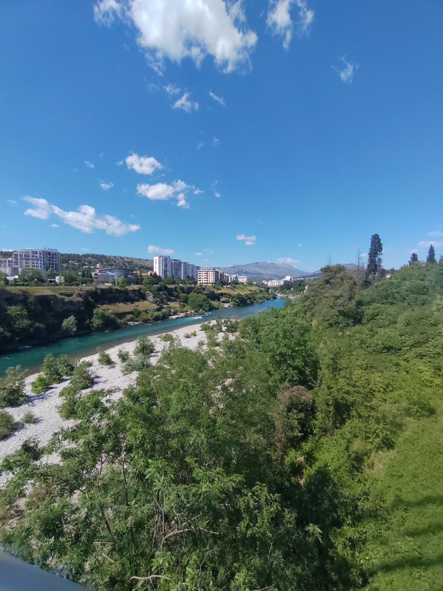 The image depicts a daytime scene in Podgorica, Montenegro. A river, likely the Morača, flows through the center of the frame, with a wide, rocky riverbed visible alongside the water. On the left bank are modern buildings, indicating an urban area, possibly part of the city center. Distant mountains are visible on the horizon. The right bank is densely vegetated with trees and shrubs, with some grassy slopes. The weather is sunny with scattered clouds in a blue sky. No people are immediately visible.