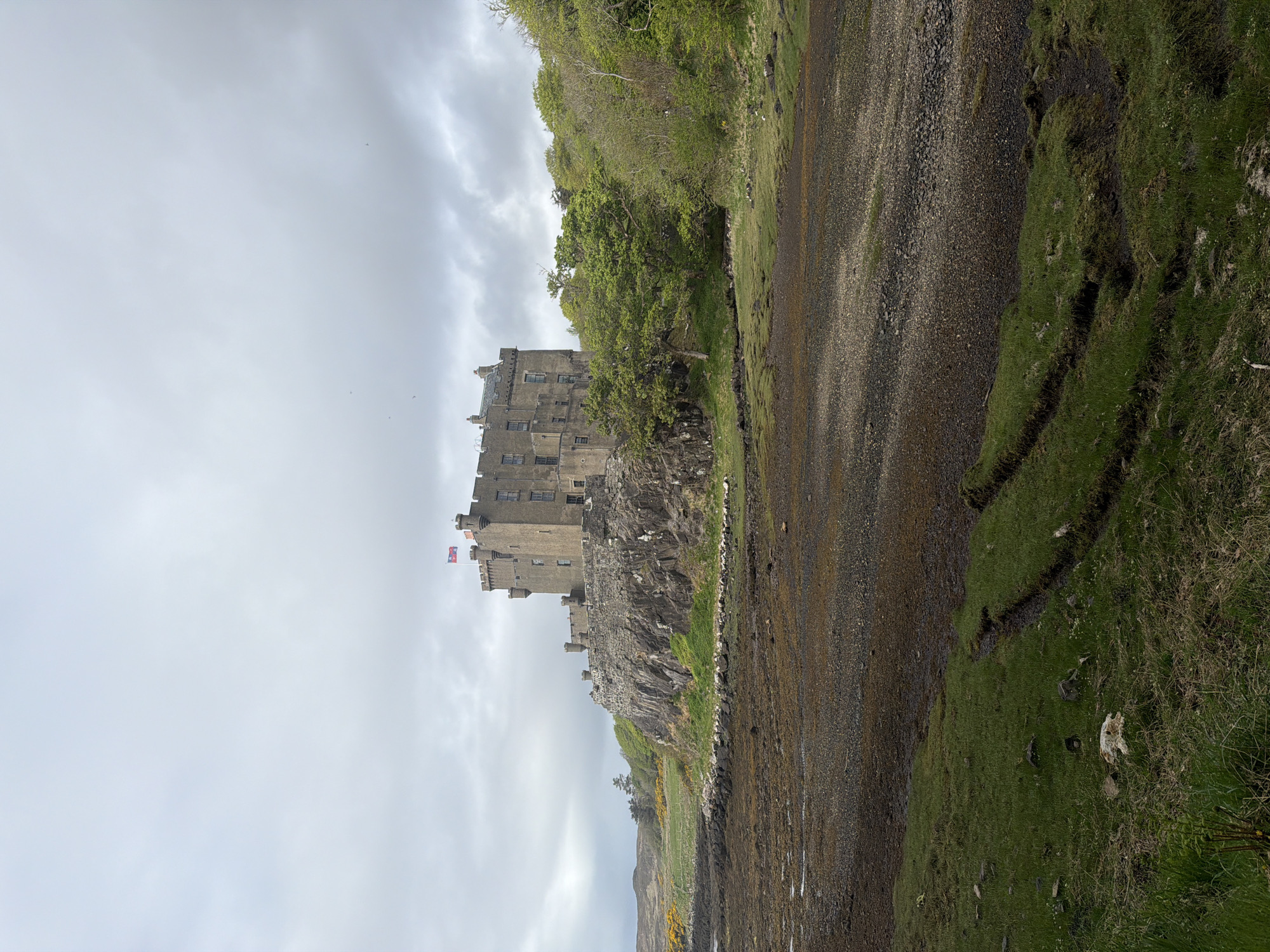 The image depicts Dunvegan Castle in Dunvegan, United Kingdom, positioned atop a rocky outcrop on the coastline. The castle, constructed of gray stone, features multiple stories, towers, and windows. Two flags are visible atop the castle. The sky is overcast with gray clouds. The foreground consists of a rocky shoreline with seaweed and green grass. A strip of land covered in rocks, dirt and grass is also visible. The perspective of the image is landscape view but tilted to be in portrait position.