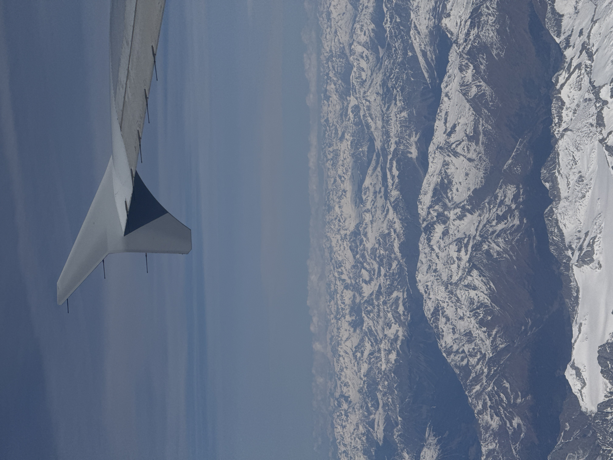 The image shows a view from an airplane window. The left side of the frame includes a portion of the airplane's wing. The right side displays a mountain range covered in snow. In between the wing and mountains, there is a hazy sky. The location is not identified in this image.