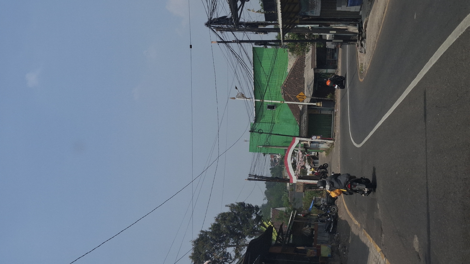 The image depicts a street scene in Cilaku, Indonesia, under a clear blue sky. A paved road curves through the frame, with a white line marking the center. Several motorcycles are present on the road and parked along the side.
In the foreground, a person wearing a dark jacket and a yellow backpack rides a motorcycle away from the camera, heading towards the curve in the road. Another motorcycle is visible further down the road, turning right. On the left side of the road, a row of buildings with dark roofs and weathered facades line the street. One building has a blue and white sign. Further back, a large, bright green building stands out, with a smaller, tiled-roof structure in front of it. A red and white archway, possibly an entrance, is visible between the buildings.
On the right side of the road, more structures with corrugated metal roofs are present. A yellow road sign with black arrows indicating a split in the road is mounted on a pole. The scene is characterized by a mix of residential and commercial buildings, with an abundance of overhead electrical wires crisscrossing the sky. The overall impression is that of a typical roadside environment in a populated area of Indonesia.