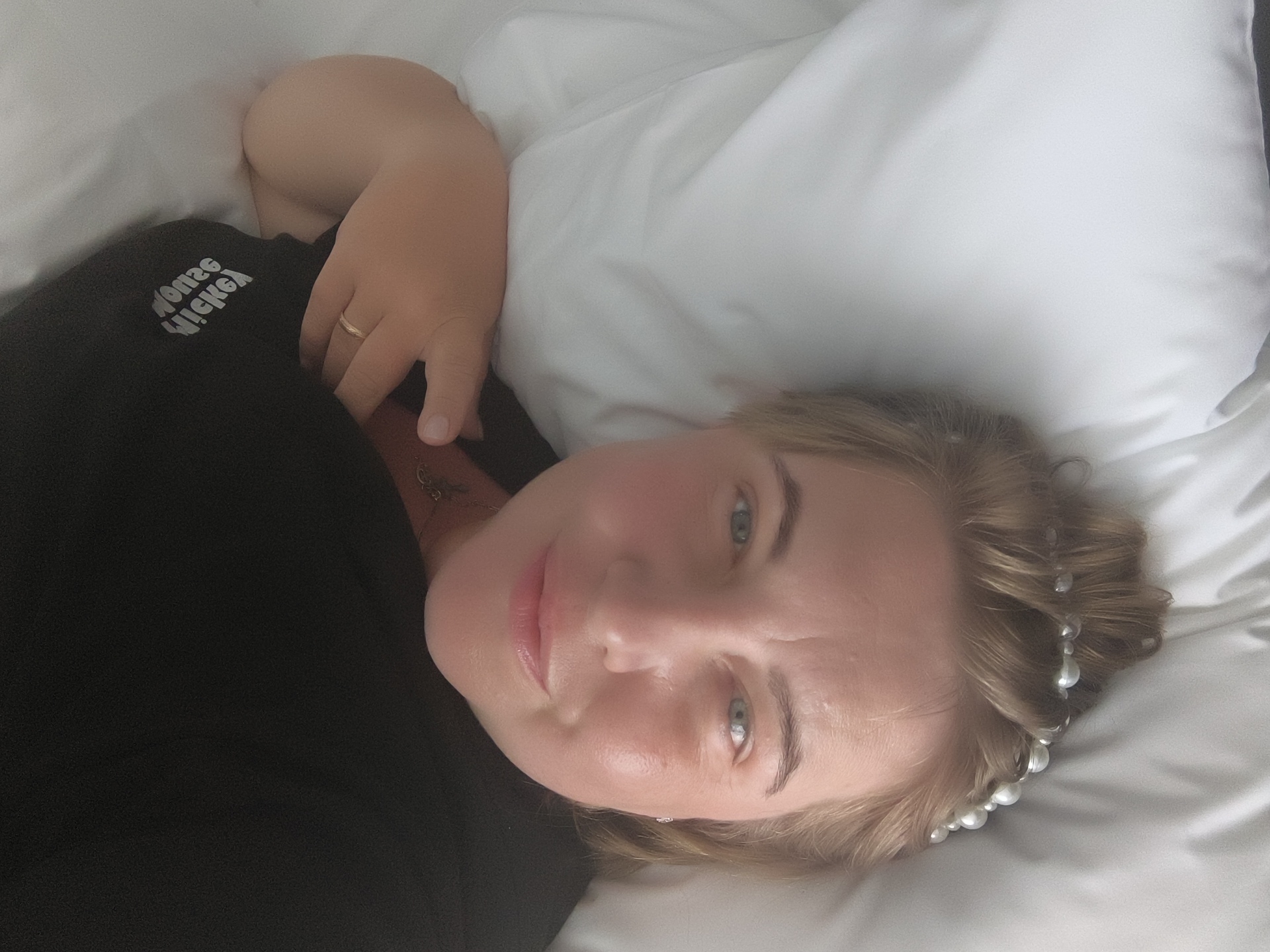 A woman with blonde hair and blue eyes is lying on a white bed, looking at the camera. She is wearing a black t-shirt with a white graphic on the left side. Her left arm is bent and resting on a white pillow. A ring is visible on her left ring finger. Pearls are woven into her hair. The setting appears to be a bedroom. There are no specific indicators of the location being in Phu Thuy Ward, Vietnam.