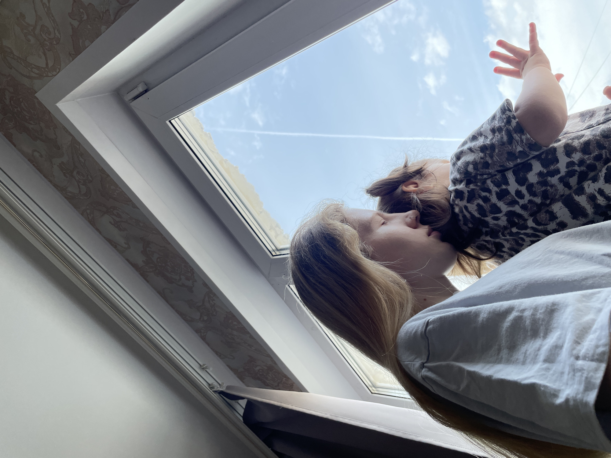 A low-angle shot depicts a woman and a child near a skylight. The woman has long blonde hair and is wearing a light-colored shirt. A small child with brown hair and wearing a leopard-print shirt is positioned above her, with one hand raised towards the sky. The open skylight reveals a blue sky with contrails. The surrounding interior includes wallpaper with a floral pattern. The location is presumed to be in Izhevsk, Russia, based on external information.