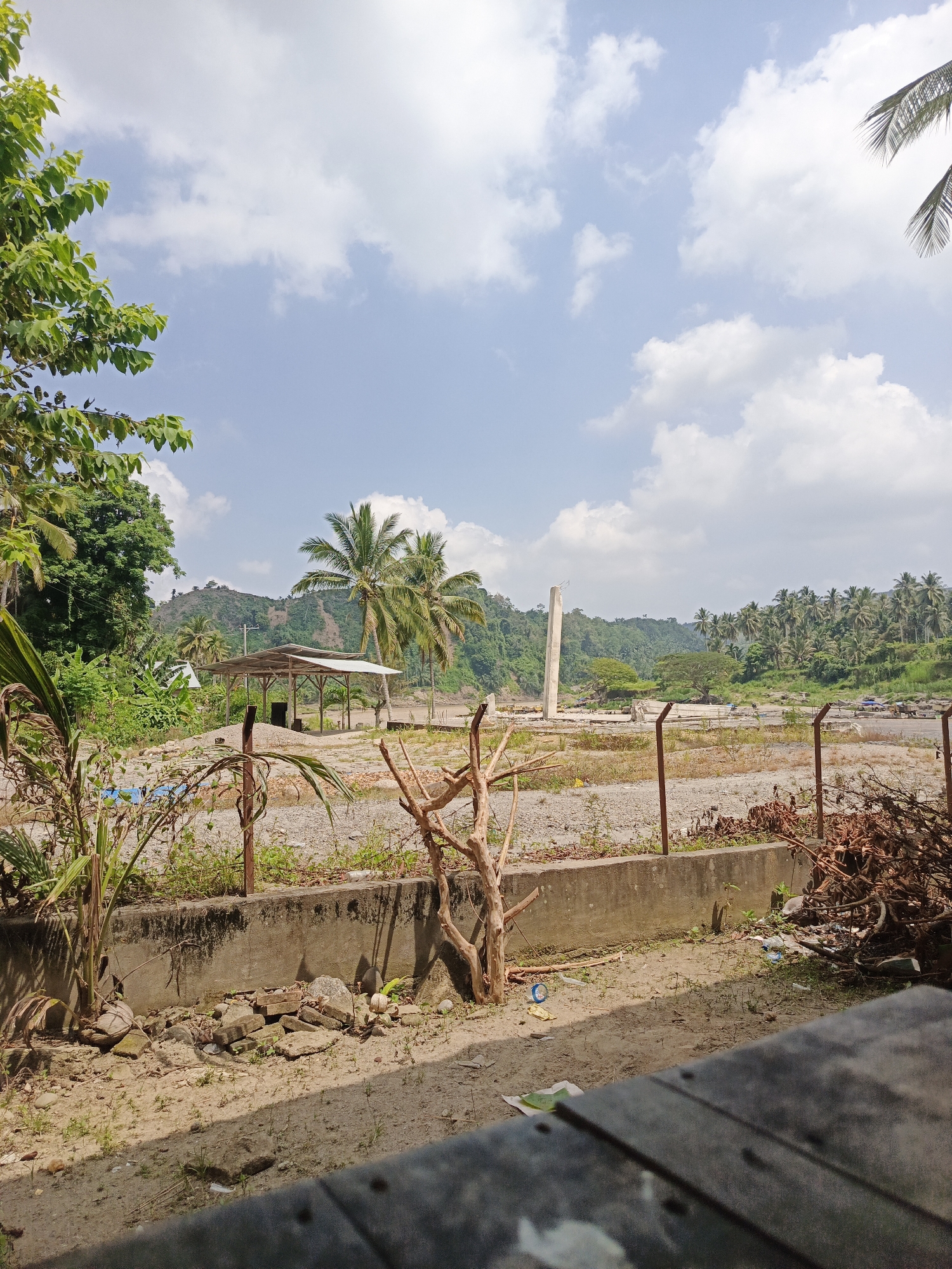 This image presents a broad outdoor view of a rural or semi-developed landscape in Simpangjaya, Indonesia, under a bright, partly cloudy sky.
In the immediate foreground, a weathered concrete ledge or barrier runs horizontally, with dry, sandy earth in front of it. This area contains sparse, small plants, a dried-up, branchy tree, and a pile of brown, dead brush and debris to the right. Some litter, including a blue plastic object and a white wrapper, is visible on the ground. Beyond the concrete barrier, a simple fence with concrete posts and wire stretches across the middle ground.
The mid-ground consists of a large, flat, unpaved expanse, likely gravel or sand, suggesting a cleared or construction area. To the left, a basic open-sided shelter with a light-colored corrugated metal roof stands among green foliage and several palm trees. Further into the mid-ground, a prominent, tall, pale concrete pillar rises vertically, hinting at ongoing construction or a remnant structure. Other smaller piles of materials or earth are scattered across this open space.
The background features lush, undulating green hills densely covered with a variety of trees, including many characteristic palm trees. The sky is a clear blue, adorned with scattered white cumulus clouds. The time of day appears to be sunny mid-day, indicated by the bright illumination and visible shadows. No people or visible text are present in the image. The scene conveys a blend of natural tropical environment with signs of human activity or development.