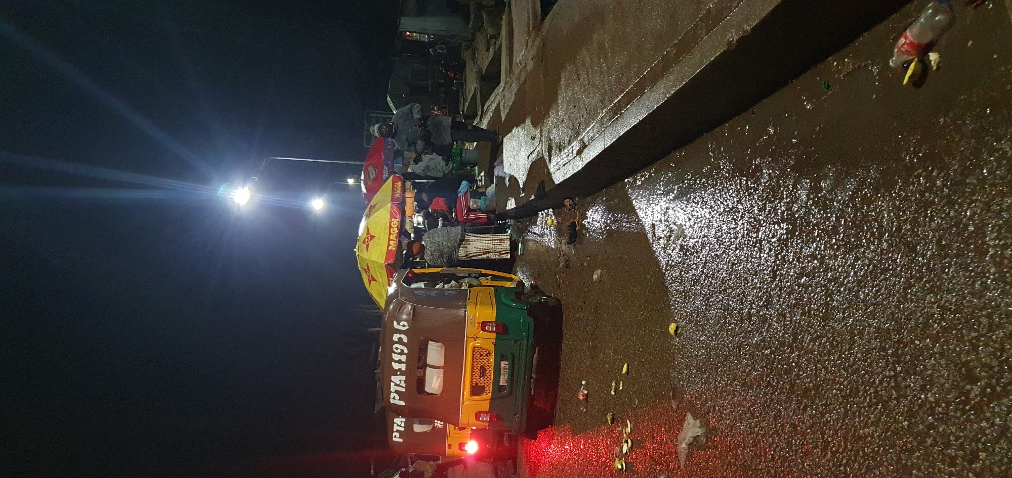 At night in Bukuru, Nigeria, an open-air market appears wet, possibly from recent rain, with puddles and reflections on the asphalt. A yellow and green auto-rickshaw, marked "PTA PTA-11936," is parked on the left, its brake lights illuminated red. In the center, a stall is set up under a large yellow umbrella advertising "MAGGI." A person wearing a patterned dress stands near the stall. To the right, a man in a gray jacket and beanie walks away from the camera. Another umbrella, red and also branded with "MAGGI," is visible behind the first. The scene is lit by overhead streetlights, which cast long shadows and create bright glare. Scattered on the wet ground are pieces of trash, including a plastic bottle and yellow fruit peels. The background is dark and indistinct, suggesting a densely populated area.