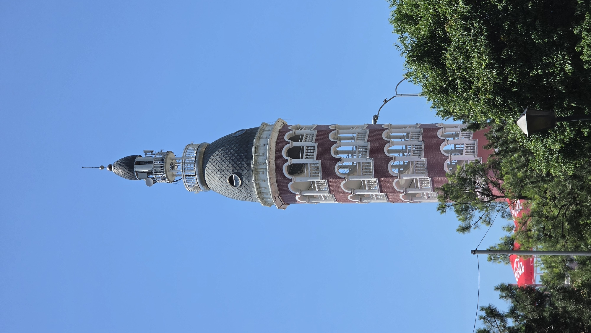 The image shows the Batumi Technological University tower, also known as the Alphabet Tower, located in Batumi, Georgia. This structure features a distinctive spiraling design with a red brick base and white colonnades. At the top, there is a dome with an observation deck and telecommunication equipment. The tower is surrounded by lush green trees and a clear blue sky. A street lamp is visible in the foreground.