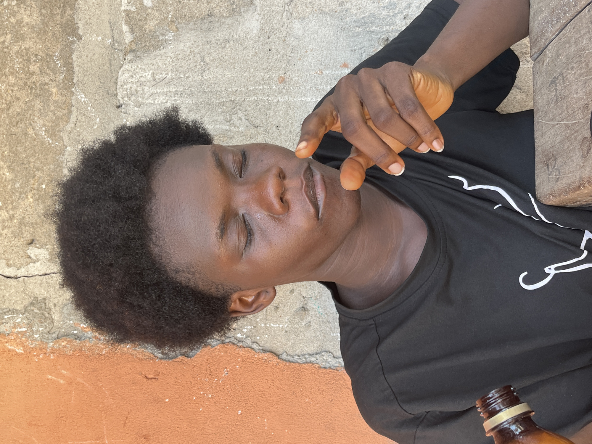 An individual with dark skin and short, dark curly hair is resting, with eyes closed, and appears to be touching their lips with their fingers. The person is wearing a black shirt with white markings and is positioned next to a rough, tan-colored wall. The bottom of a brown bottle is visible in the lower right corner of the image. The lower part of the wall is painted orange. The location is believed to be in Badagry, Nigeria.