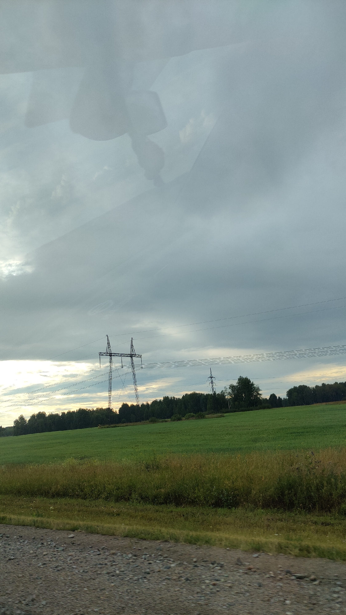 A telephoto shot captures a wide, rural landscape under a cloudy sky. In the mid-ground, two sets of tall, metallic power line towers stand against a backdrop of dense, dark green trees. The towers are connected by thick black wires, which stretch across the frame. In front of the trees is a bright green, rectangular field. The foreground is a rough, grey gravel road, with a strip of overgrown grass and weeds separating it from the field. The sky is heavily overcast with grey clouds, but there are patches of lighter sky near the horizon, suggesting it might be dawn or dusk. A faint reflection, possibly from a car window, is visible at the top of the image.