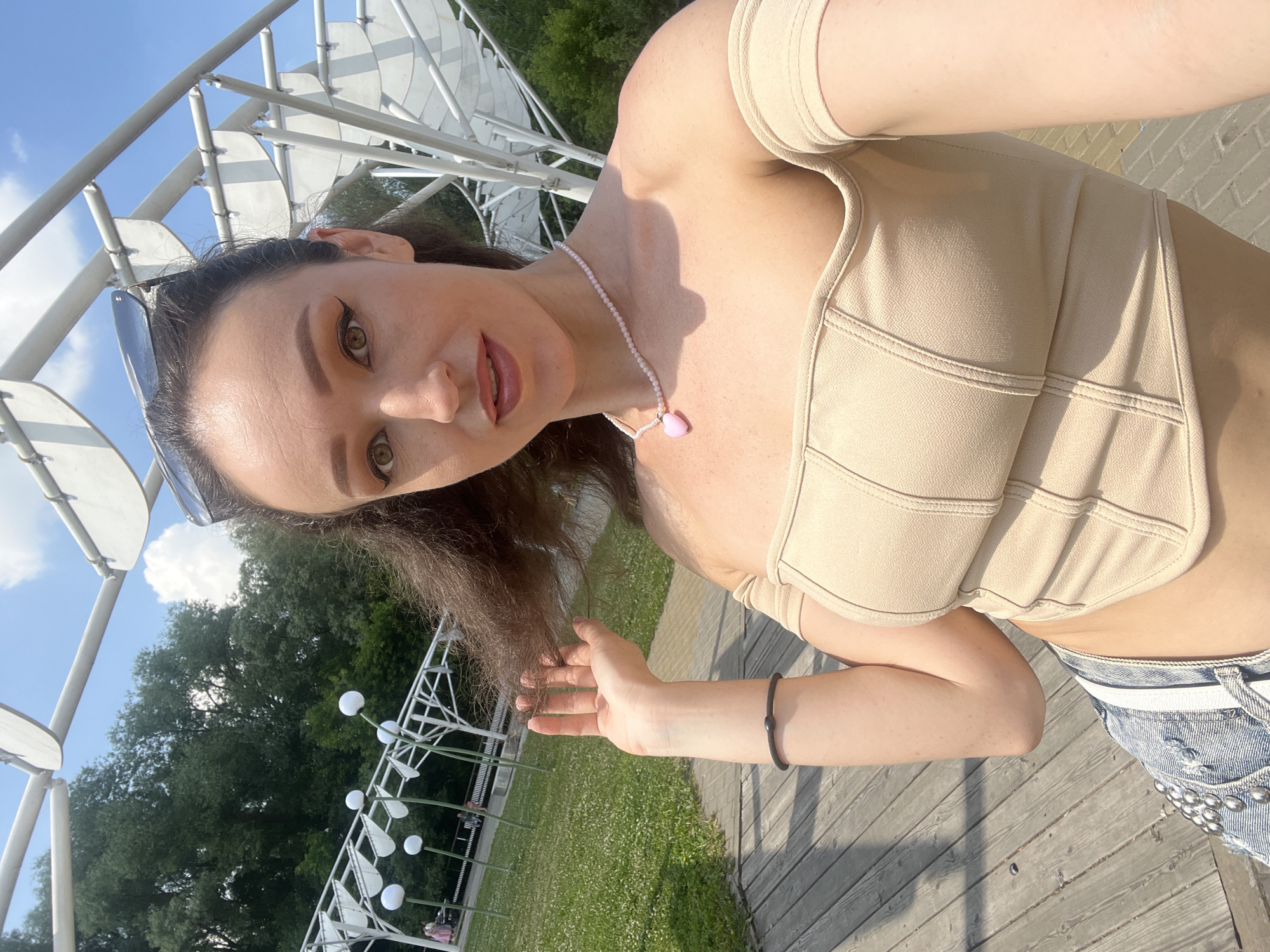 A woman is taking a selfie in an outdoor public park, possibly in Kazan, Russia. She is seated on a wooden deck, with a metal and white abstract art installation behind her. She has long, dark, wavy hair, which she is touching with her right hand. She is wearing a beige, off-the-shoulder corset-style top and distressed denim shorts with embellishments. A pink heart pendant on a pearl necklace is visible. Her left arm rests on her leg, and she is wearing a dark bracelet. In the background, there are trees, grass, and other park visitors. The sky is blue with scattered white clouds.