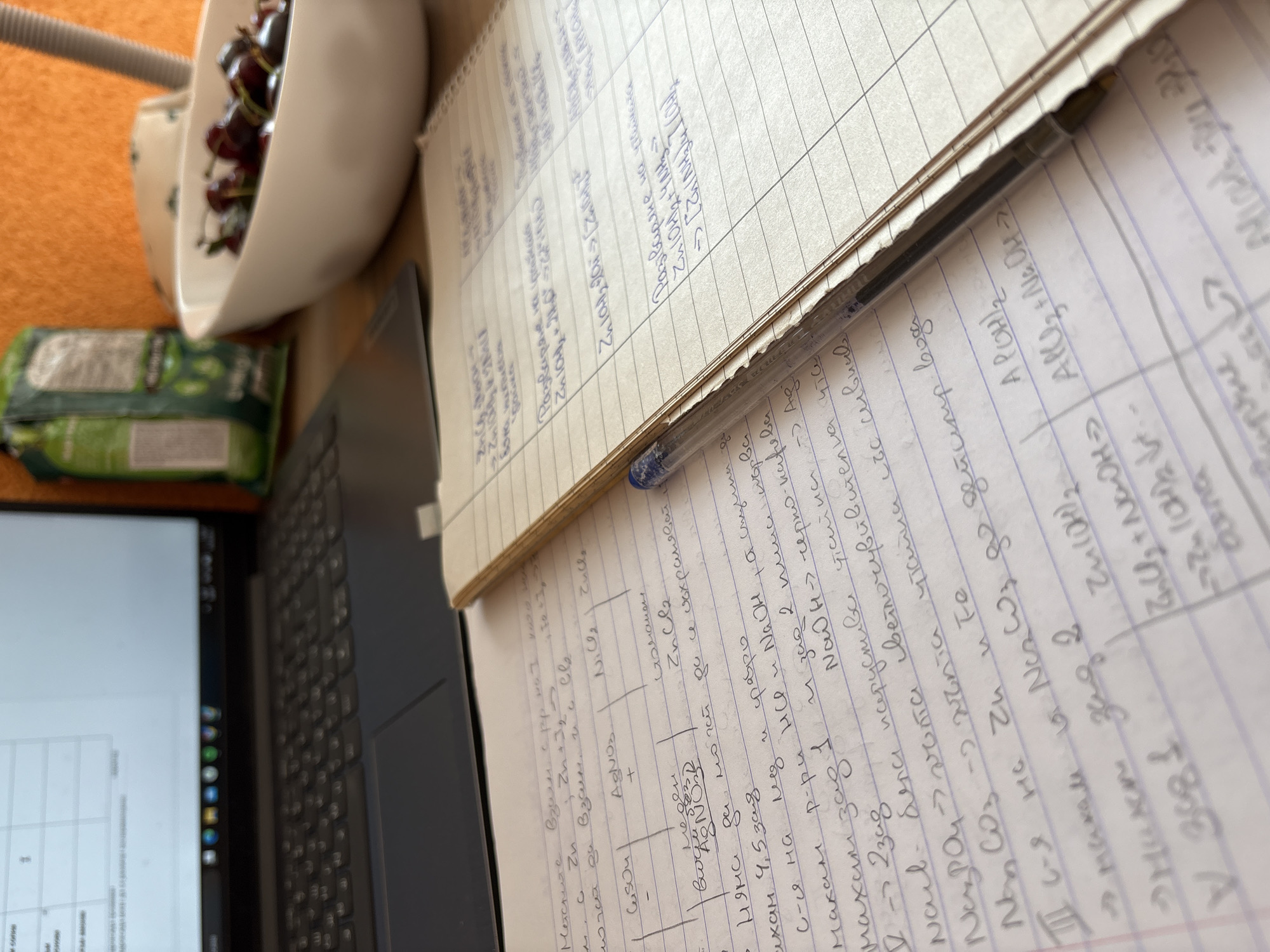 The image depicts a close-up, eye-level view of a desk with various objects related to studying and a bowl of cherries. The dominant elements are a notebook with handwritten notes and chemical equations, a laptop partially visible on the left, and a white bowl filled with dark red cherries on the right. The notebook is open, and a blue pen rests on its pages, poised over the writing. The notes appear to be in Bulgarian and relate to chemistry, with chemical formulas and reactions clearly legible in places. The environment is indoor, likely a home or study space, with an orange textured background suggesting a wall or fabric. The lighting is natural and appears to be coming from the right, casting soft shadows. The overall scene suggests a moment of study or work, interspersed with a snack. There are no people visible in the image. The location of Panagyurishte, Bulgaria is not discernible from the visual information provided in the image.