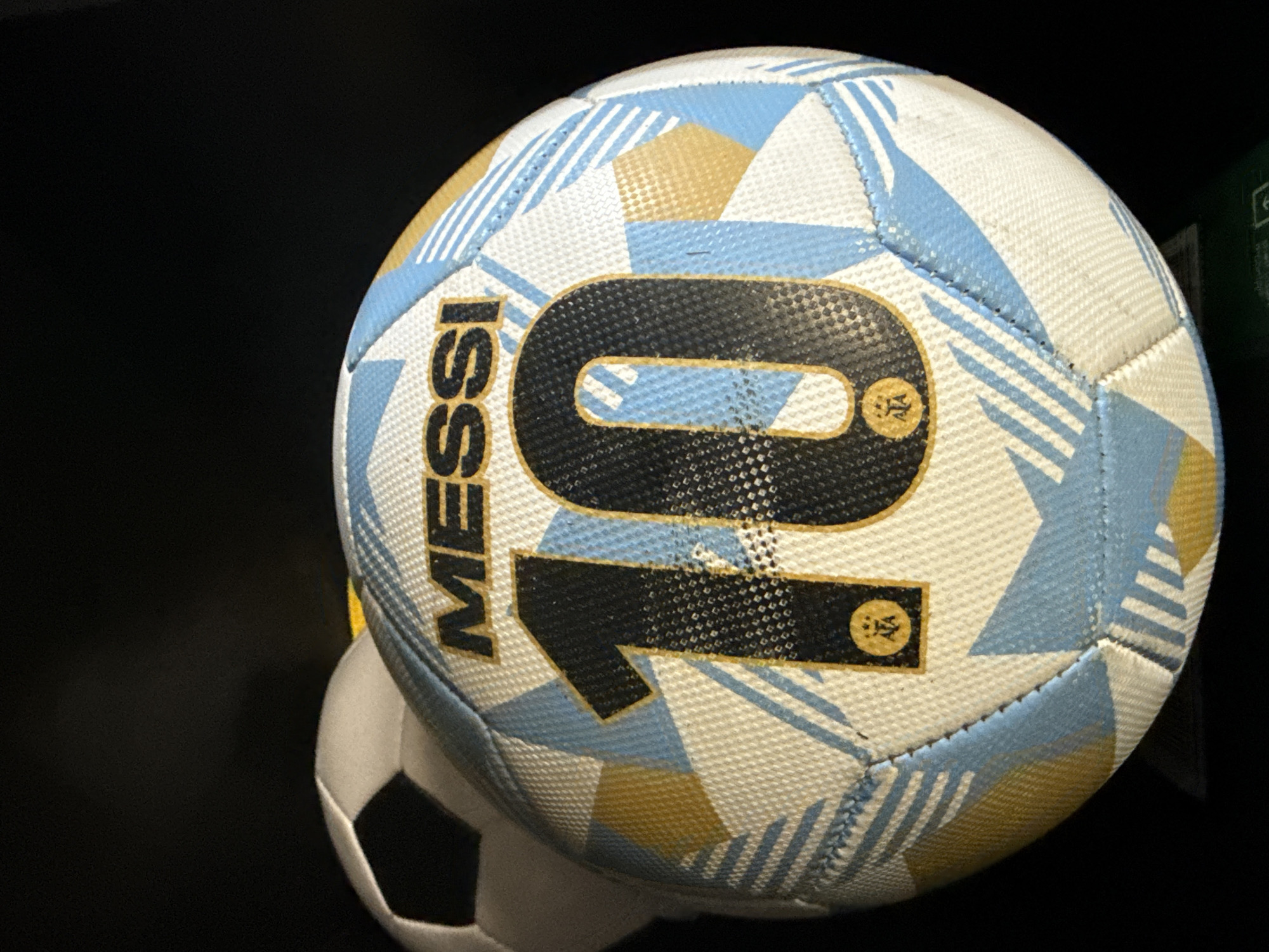 A close-up shows a soccer ball with "MESSI 10" printed on its surface, alongside a second, partially visible soccer ball in the background. The main ball is patterned with light blue and gold geometric shapes on a white base. The number "10" is in a large, dark font with small circular patches near the top and bottom of the numeral. The background is dark, suggesting an indoor setting. No people are visible. The location in Davie, United States of America is not visually apparent.
