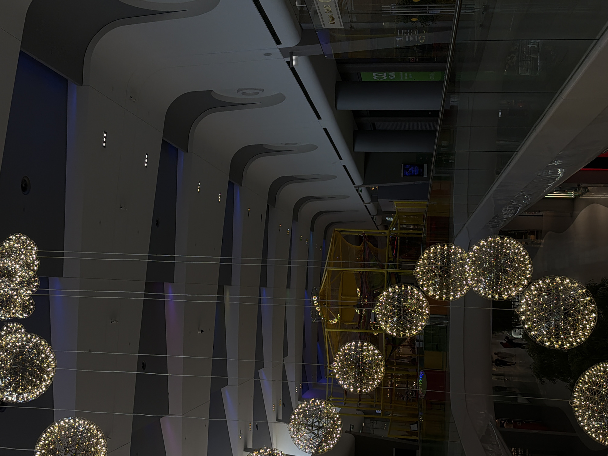 The image depicts the interior of the OZ Mall in Krasnodar, Russia. The scene is dominated by a series of spherical, illuminated light fixtures hanging from the ceiling by thin wires, creating a decorative canopy. Below these lights, a multi-level interior of a shopping mall is visible. On the upper level, a large, brightly colored indoor playground structure, primarily yellow, is present. A glass railing lines the upper walkway, and further into the mall, shops are visible along the walls, some with illuminated signs. People are seen walking in the distance on the lower level of the mall. The architectural style of the mall is modern, with curved ceilings and contrasting dark and light surfaces. The lighting in the scene is a mix of ambient light from the mall's infrastructure and the prominent decorative lights.