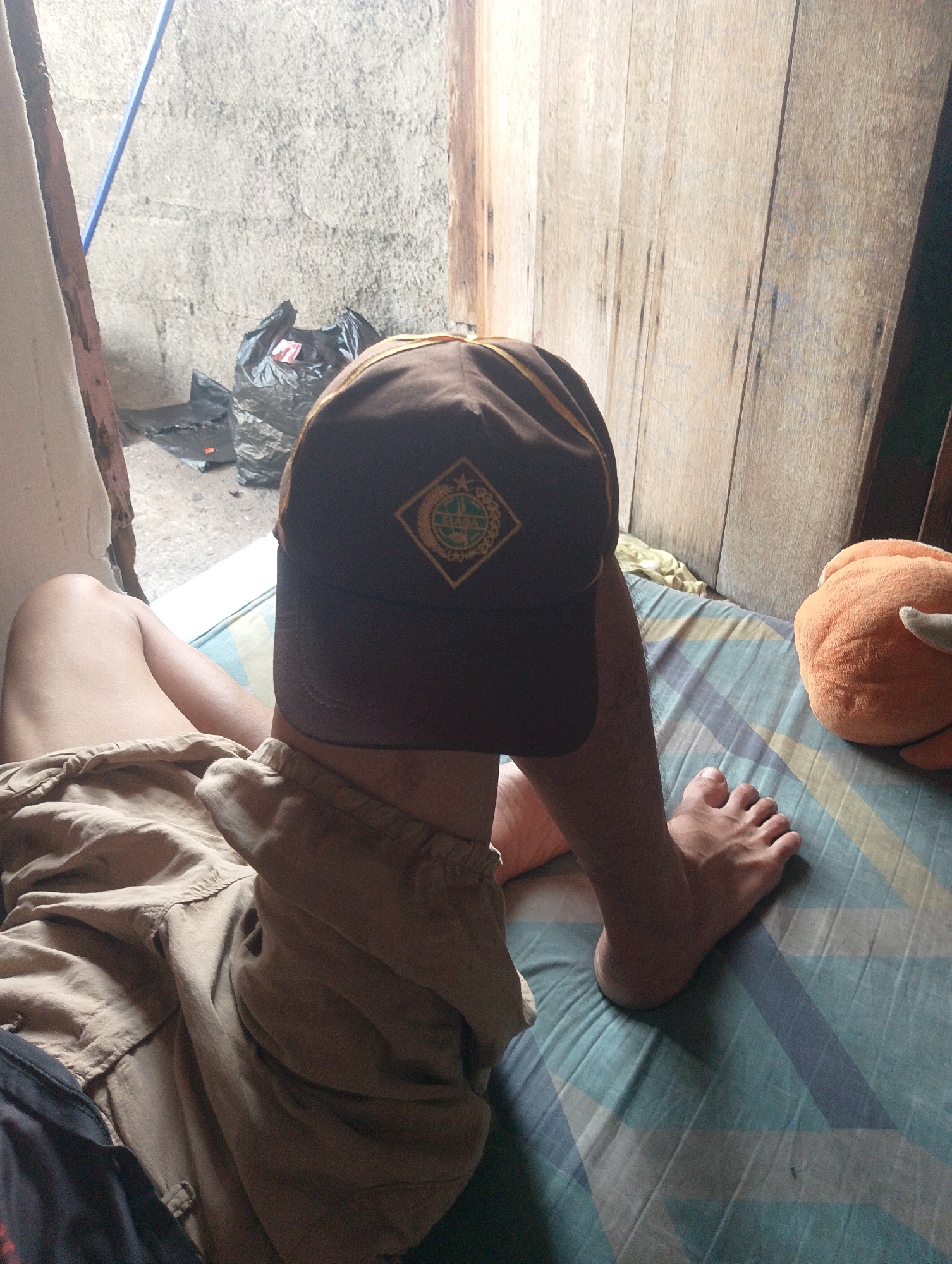 The image depicts a person, whose face is obscured by a brown baseball cap, resting on a bed. The setting appears to be an indoor space with rustic elements. Behind the person, a rough, gray concrete wall is visible, with a blue pole leaning against it. A black plastic bag is also present near the wall. To the right of the person, a wooden wall with vertical planks creates a rustic backdrop.
The person is wearing khaki shorts and is lying on a bed with a blue, green, and yellow striped mattress cover. The person's legs and feet are visible. On the bed to the right of the person, an orange, fuzzy spherical object, possibly a stuffed toy, is also present. The lighting suggests it is daytime, with natural light coming from an unseen source. The overall atmosphere seems relaxed and informal. There is a small, circular emblem on the baseball cap, but the text on it is illegible. The location context is South Jakarta, Indonesia.