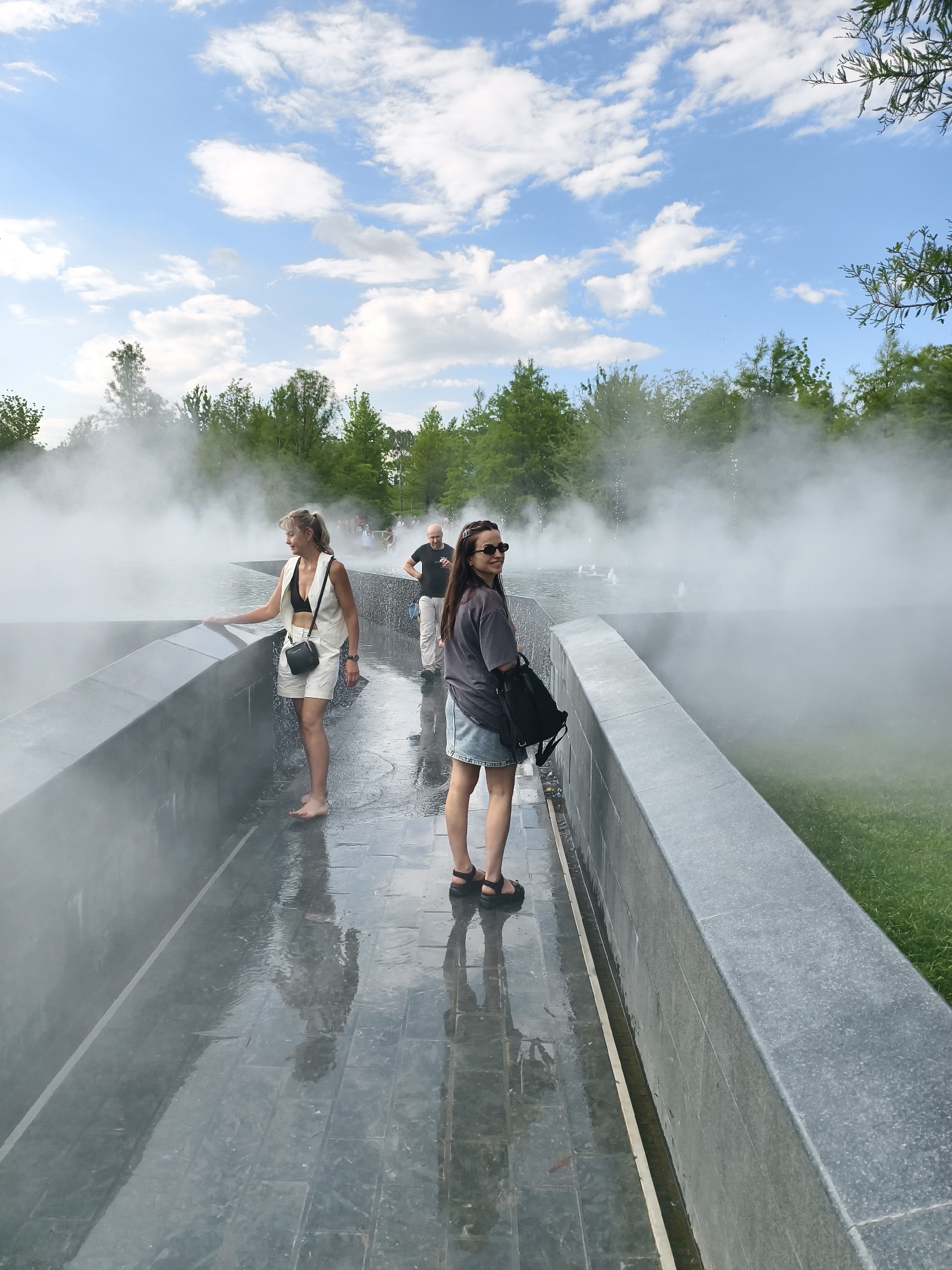 The image depicts a shallow water feature with mist rising from it, creating an atmospheric effect. Two women are present in the foreground. The woman on the left is facing away from the camera, wearing a white vest, a black bikini top, and white shorts. She is barefoot and touching the edge of the water feature. The woman on the right is facing the camera, wearing sunglasses, a dark t-shirt, and a denim skirt. She has a backpack slung over her shoulder. A man in a dark t-shirt and light pants is visible in the background, walking away from the camera. The area surrounding the water feature is paved with dark tiles that are wet and reflective. Green grass is visible to the right of the paved area, and trees form a backdrop. The sky is blue with scattered white clouds.
This location is the "Fountain of the Sun" in the "Galitsky Park" in Krasnodar, Russia.
