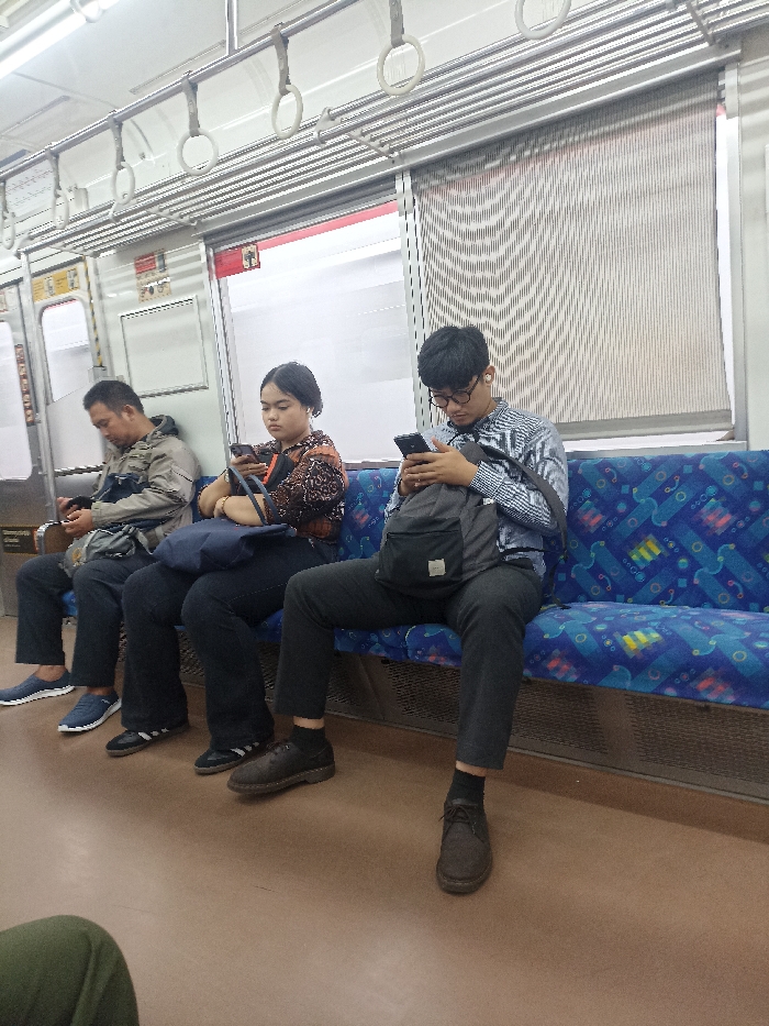 Three individuals are seated on blue and white patterned train seats facing the right side of the image. The man on the right is wearing a light blue and white striped shirt, blue jeans, a black backpack, and black shoes. He is looking down at his phone in his right hand. The woman in the middle is wearing a brown and white patterned blouse, black pants, and black shoes. She is looking at her phone in her left hand. The man on the left is wearing a blue jacket, dark blue jeans, and black shoes. He is looking down at his phone in his right hand. They are all on a train in Bojong Gede, Indonesia (latitude: -6.501, longitude: 106.796). The interior of the train is lined with a light grey material and decorated with silver metal handles. There are two windows in the background. The train appears to be empty besides these three individuals.