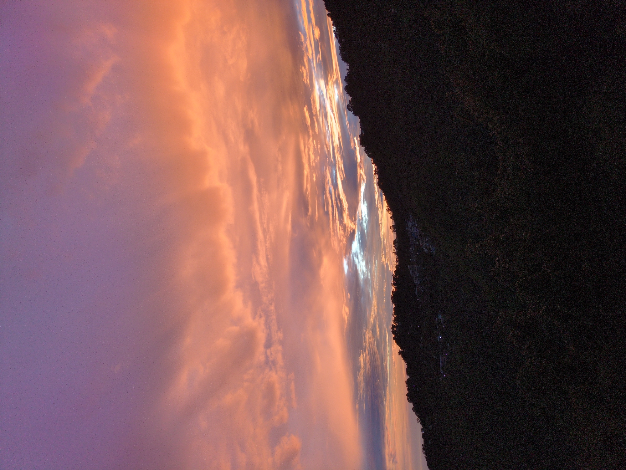 A wide shot captures the sky over a dark, tree-covered mountainside dotted with buildings in Dharamkot, India. The sky is painted with a gradient of colors, transitioning from a soft purple at the top to vibrant oranges and pinks in the clouds below. Thin streaks of brighter light are visible where the clouds meet the horizon. The mountainside is predominantly dark, with the silhouettes of trees and some scattered lights indicating the presence of homes or structures. The overall scene depicts a twilight or dawn setting.