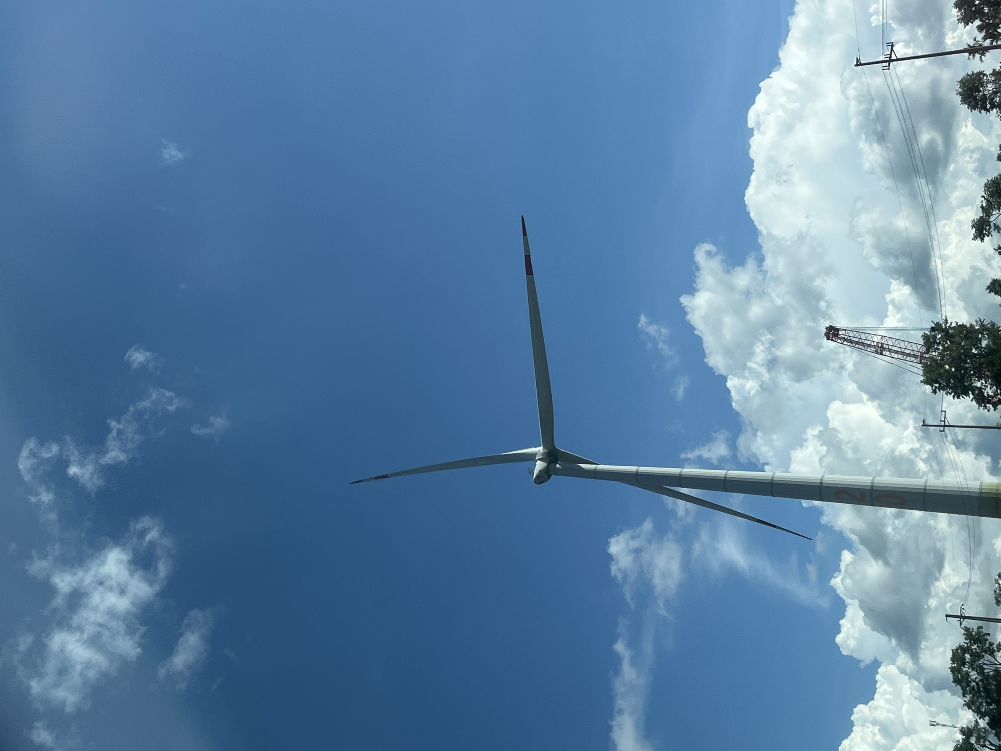 A large wind turbine stands tall against a bright blue sky dotted with white clouds. The turbine is white with two long blades, one angled upwards and the other slightly downwards. The tips of the blades have red accents. The lower section of the turbine's tower is visible, featuring orange lettering that appears to spell out "20". In the background, a red construction crane is visible above a line of trees. Power lines and poles are also present in the lower part of the image. The scene appears to be an open area, possibly a field or industrial site, given the presence of the wind turbine and crane. The overall impression is one of renewable energy infrastructure under a clear sky. This scene is located in the city of Tri C3, Vietnam.