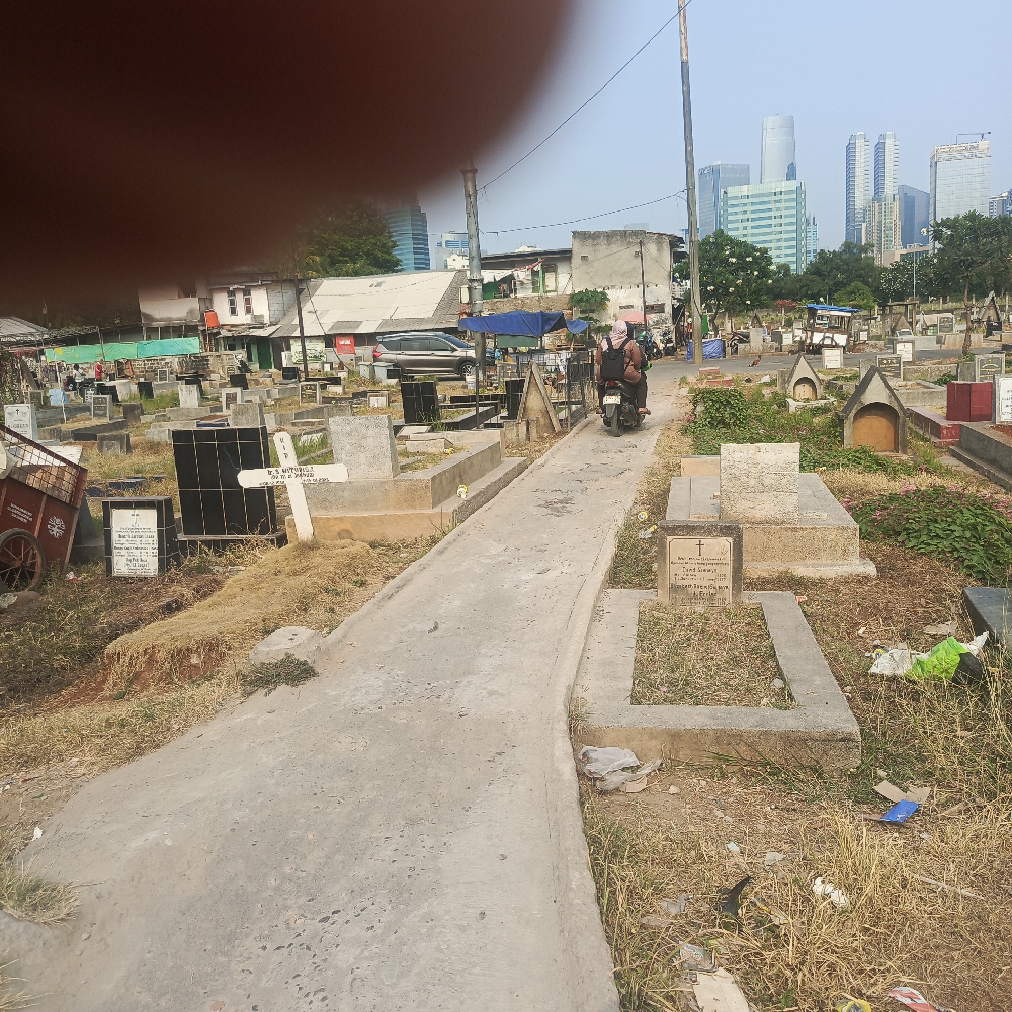 The image depicts a cemetery in South Jakarta, Indonesia. The scene is characterized by numerous gravestones and burial plots, with some featuring ornate headstones and others simple markers. A concrete pathway winds through the cemetery. In the background, modern skyscrapers rise above the treeline, indicating the proximity of an urban area. A woman and a child are seen riding a motorcycle down the path, while other people are present in the distance near some buildings. The ground is covered in dry grass and scattered debris, including plastic bags and cardboard. The sky is clear and the lighting suggests it is daytime.