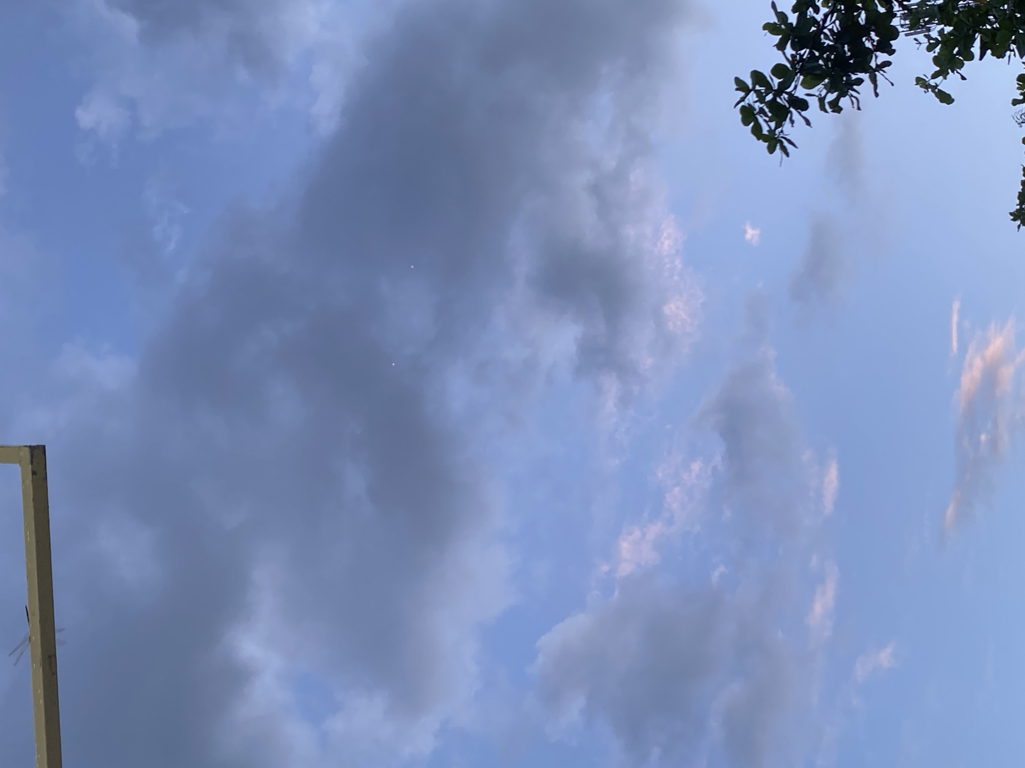 A clear, bright sky with scattered clouds, tinged with pale pink and purple, dominates the image. In the upper left, a golden-colored structure, possibly part of a building or fence, is visible, with a dark insect on its surface. The lower right corner shows the dark green leaves of a tree. Two small, bright pink, circular lights are visible in the sky, positioned slightly to the right of the center of the frame. The lighting suggests it is either dawn or dusk. The scene appears to be an outdoor setting in Gambut Barat, Indonesia.