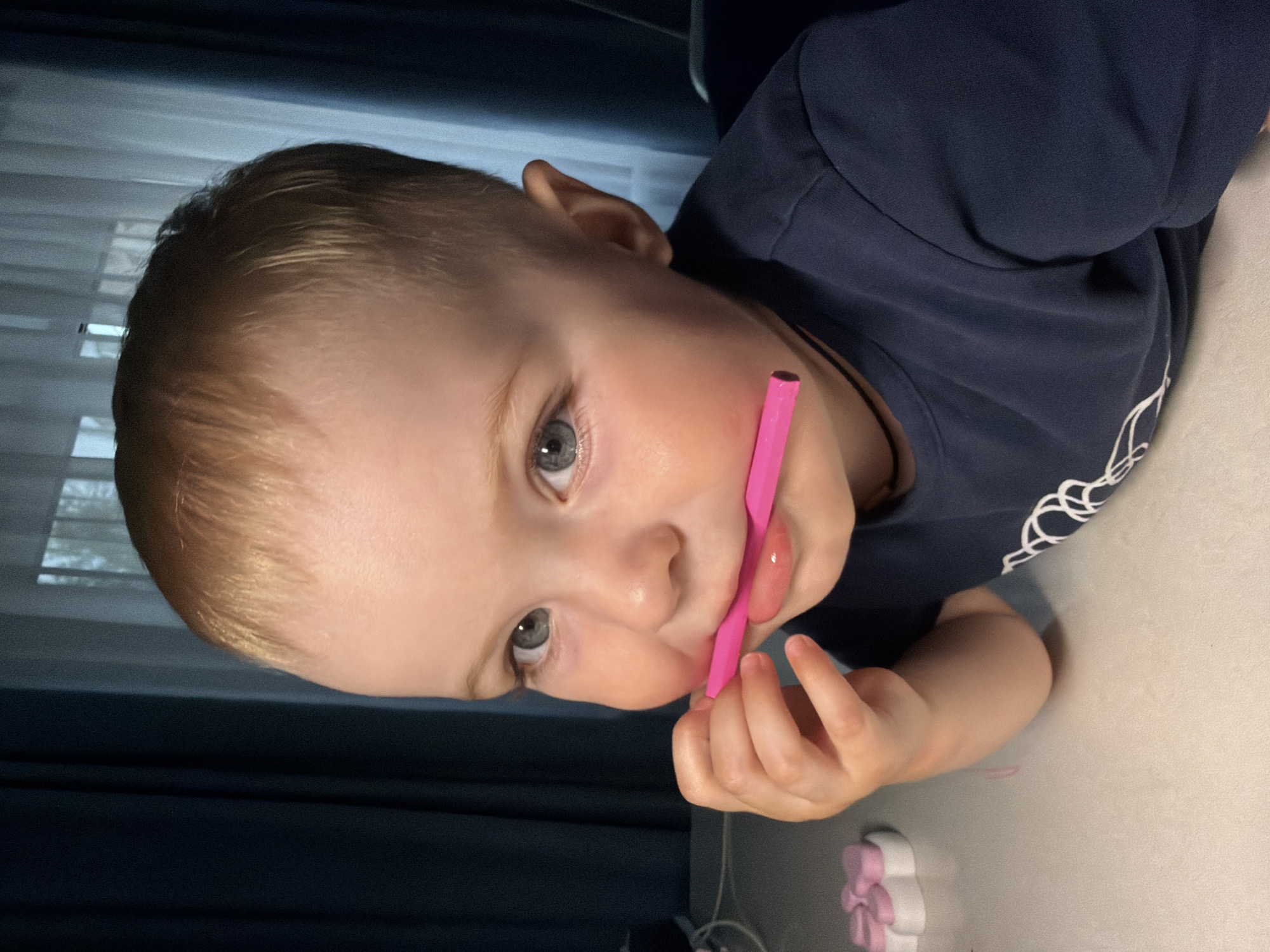 A close-up, eye-level shot shows a young Caucasian child with light brown hair, blue eyes, and fair skin. The child is wearing a dark blue t-shirt and is holding a pink object, possibly a crayon or pencil, in their mouth. The child's right hand is near their mouth, touching the object. The background consists of dark blue curtains and a window with sheer white curtains, suggesting an indoor setting. The lighting is dim. The image appears to be taken in Votkinsk, Russia.