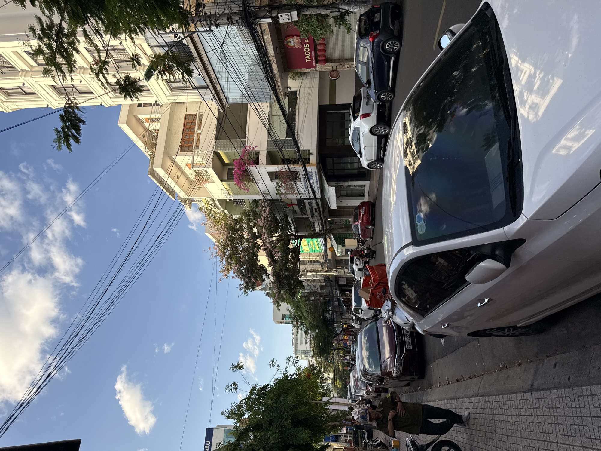 The image depicts a street scene in Nha Trang, Vietnam. A person, likely a street vendor or security guard due to their uniform, is standing on a sidewalk. Several cars are parked and driving along the street, with a prominent white car in the foreground. Buildings line the street, some with balconies adorned with plants. Electrical wires are strung across the sky, a common sight in urban Vietnam. The sky is blue with scattered white clouds, suggesting it is daytime. The overall impression is of a typical city street with ongoing traffic and commercial activity.