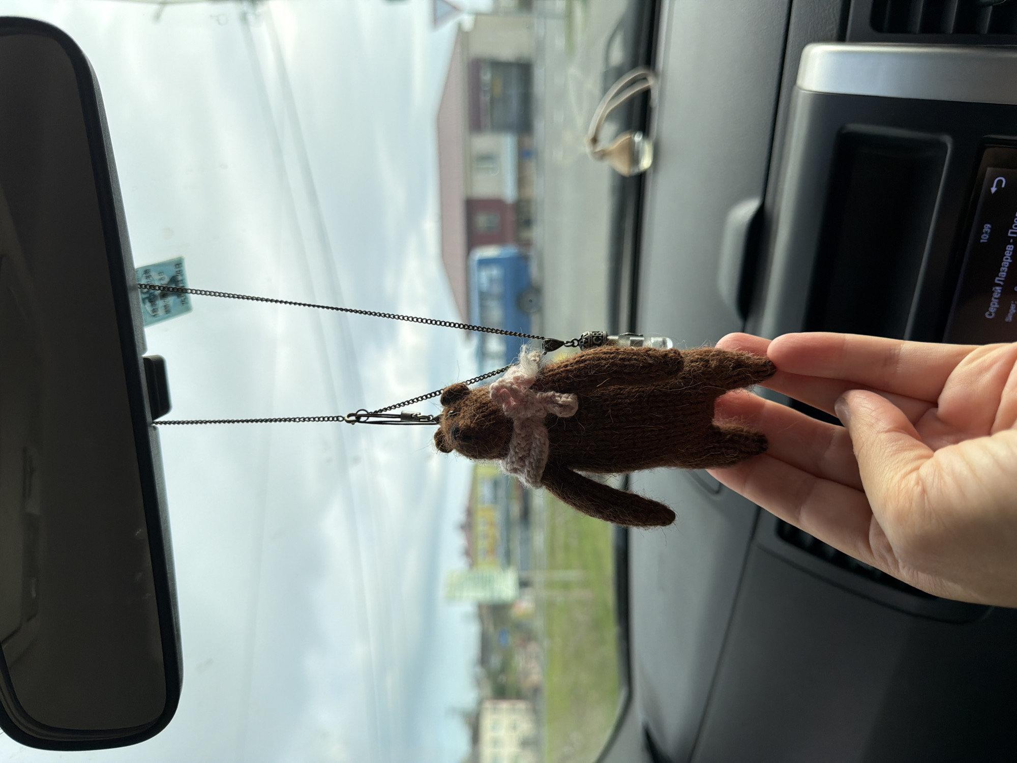 A knitted brown bear charm with a pink scarf is hanging from a car's rearview mirror. The bear is in focus, held by a person's hand in the foreground. Outside the car window, a blurred street scene shows buildings, a blue bus, and a traffic sign. The sky is overcast with clouds. The image appears to be taken from inside a vehicle in Yuzhno-Kurilsk, Russia.