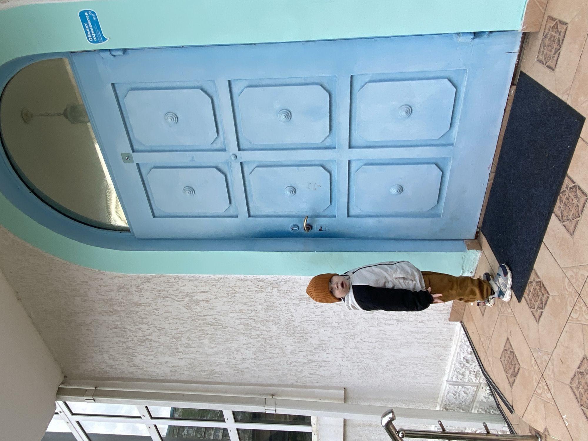 A young boy stands outside a blue door with a semicircular window in Sochi, Russia. The boy wears a brown hat, a white and black jacket, and brown pants. A dark blue mat is on the floor next to the door, which is set in a light blue archway. The floor tiles are beige with a brown diamond pattern. A window is visible in the lower left of the image. A blue sticker is visible on the upper left of the door.