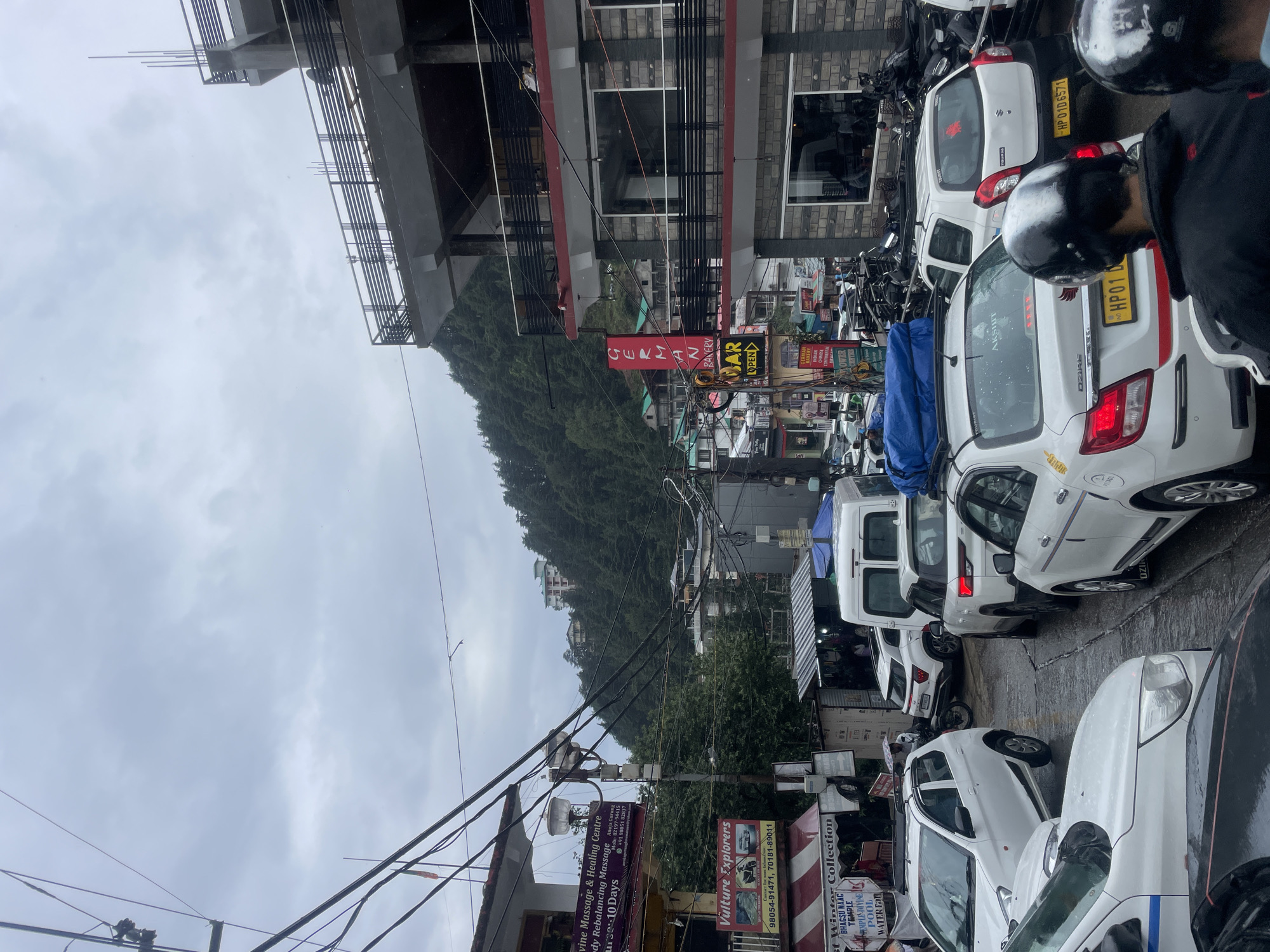 The image depicts a congested street scene in Bhagsu, India, characterized by a traffic jam and dense urban development. Several white cars, including a Maruti Suzuki Alto and a Swift Dzire, are stopped in traffic. A white van is also present. The road appears wet, suggesting recent rain.
Buildings line the street, some with multiple stories and balconies. Signs for various businesses are visible, including "Vulture Explorers," "Bhagsu NAAC Temple," and "German Bakery." A prominent red sign with white lettering reads "GERMAN BAKERY." A vertical yellow sign with red lettering indicates "BAR OPEN."
The background shows a steep, tree-covered hillside with more buildings nestled among the trees. Overhead, a complex network of electrical wires crisscrosses the sky, which is overcast and cloudy. Two individuals wearing helmets are visible in the foreground, likely on motorcycles or scooters, also caught in the traffic. The overall impression is one of a bustling, somewhat chaotic street environment in a hilly region.