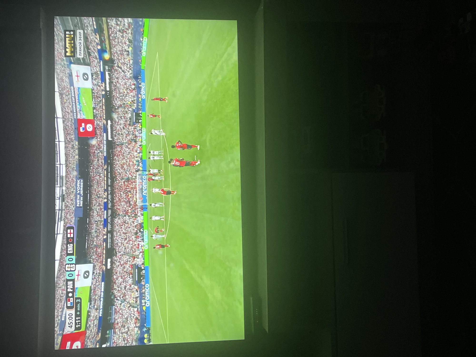 The image shows a large television screen displaying a soccer match. The score is Panama 0, England 0, with the game in the 45th minute plus 3 minutes of added time. The match is identified as being part of the FIFA World Cup 2026 and is taking place in New York, New Jersey. The stadium is full of spectators. There are players on the field wearing red and white uniforms. The screen is positioned in a dark room, with two toy cars visible in the lower right portion of the image, suggesting it is a home entertainment setup. The location is stated as Шебунино, Russia, which is inconsistent with the on-screen location of New York, New Jersey.