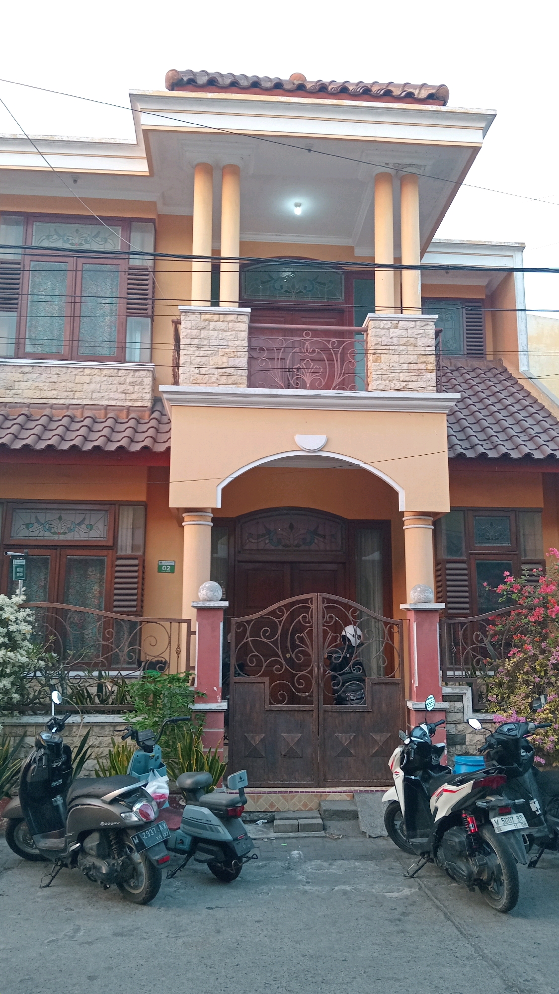 In the city of Manyarsidomukti, Indonesia, a two-story house with an orange facade is captured. The house features a red tile roof, large windows with wooden frames, and decorative ironwork on the balconies and gate. Two scooters are parked in front of the house, one black and one gray, both facing left. The gate is made of dark brown iron with intricate scroll designs. In the foreground, the concrete ground is visible, with some greenery and flowering bushes to the left and right of the entrance. The sky above is overcast and white.