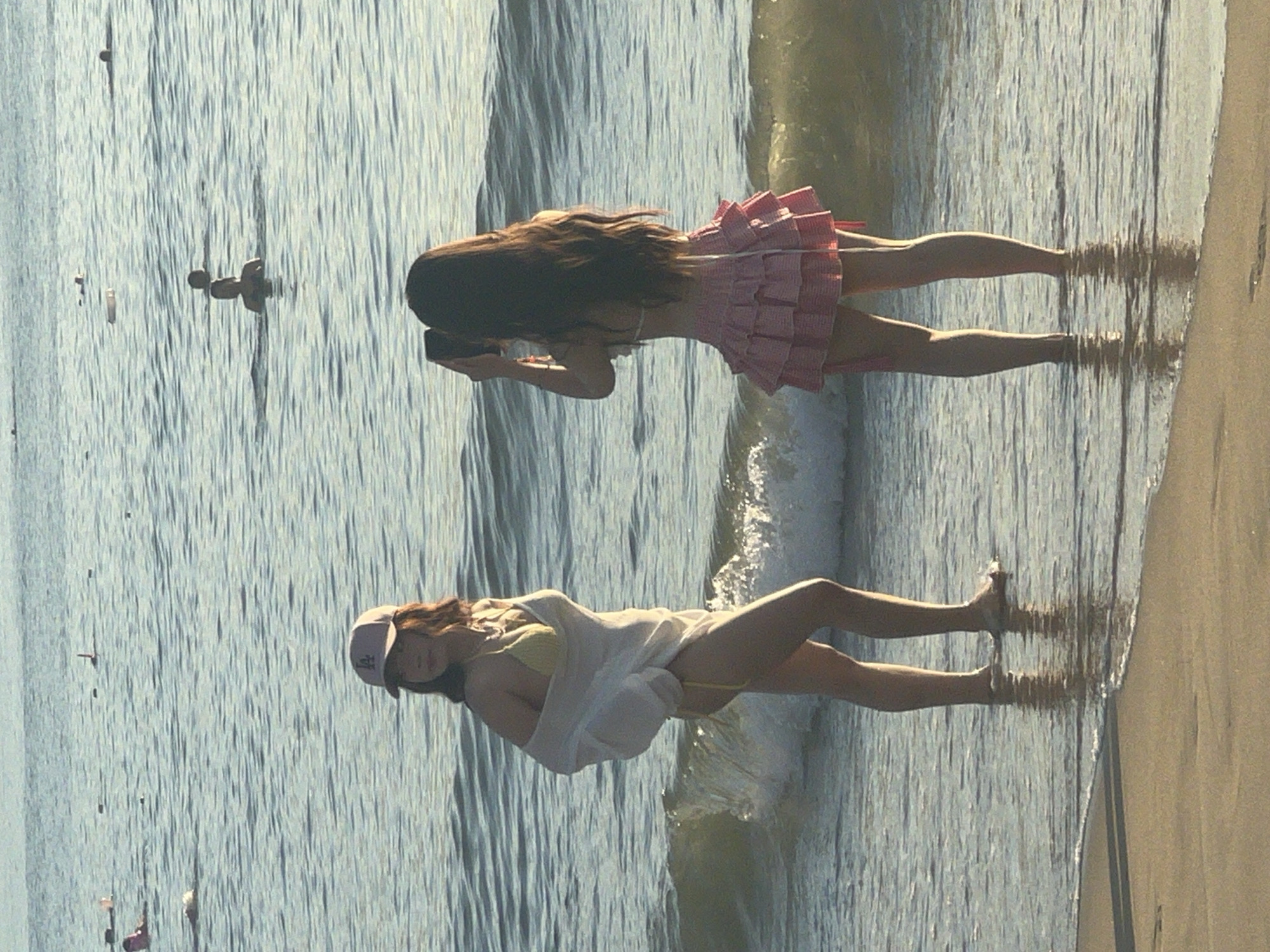 The image depicts a beach scene at sunset in Da Nang, Vietnam. In the foreground, two young women are standing in the shallow water. The woman on the left, facing away from the camera and towards the sea, is wearing a yellow bikini and a sheer white cover-up. She has a pink baseball cap on her head. The woman on the right is facing the sea and appears to be taking a photograph with her phone. She is wearing a white bikini top and a pink ruffled skirt. The sea behind them is calm with small waves, and there are several other people swimming in the distance. The sand of the beach is visible in the bottom part of the frame. The overall lighting suggests it is late afternoon or early evening.