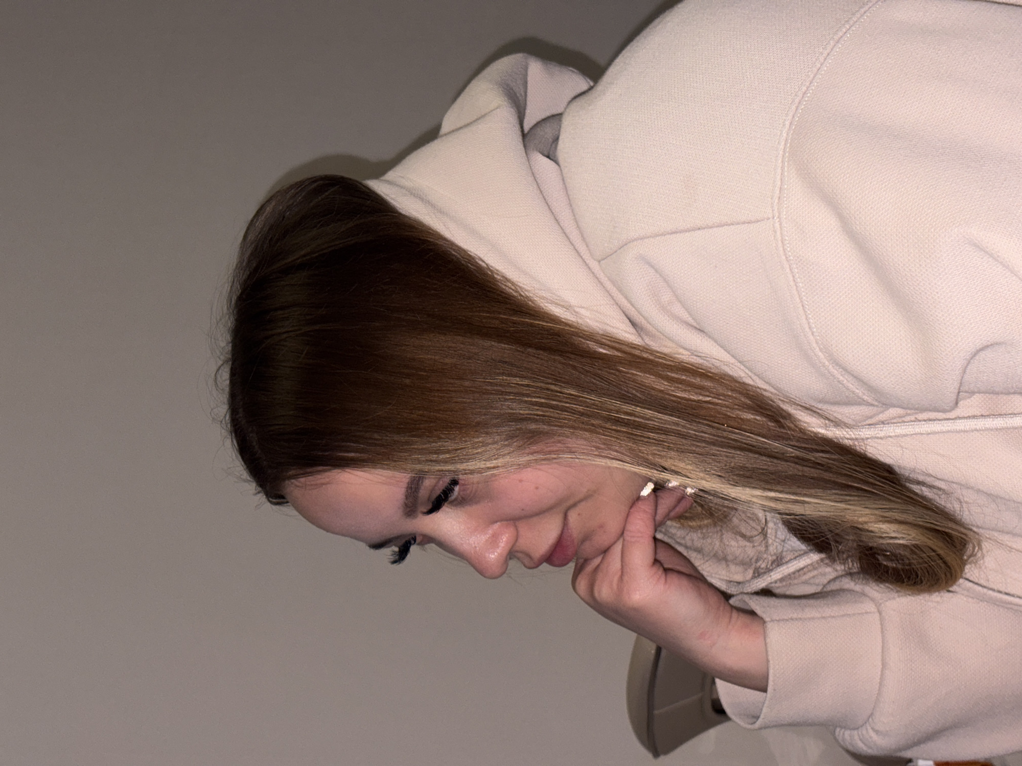 A young woman with long, brown-to-blonde ombré hair is resting her chin on her hand, looking down. She is wearing a light pink hooded sweatshirt. The background is a plain, light gray wall. The location is not discernible from the image.