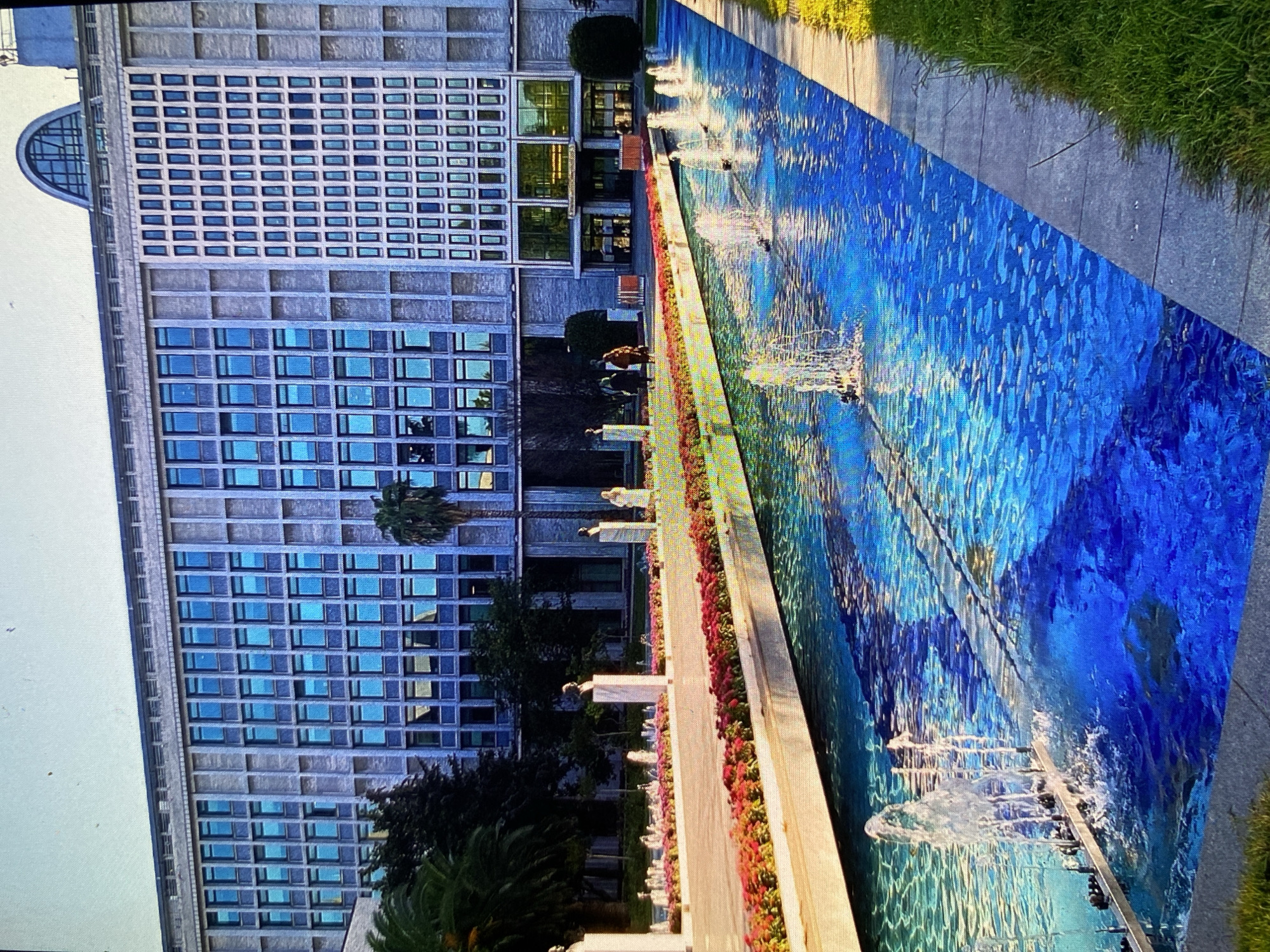The image captures a vibrant, tilted view of a long, linear water feature running parallel to a large, modern building, likely situated in Bankya, Bulgaria.
The central subject is an elongated fountain with strikingly blue water, suggesting a painted basin or tiled bottom, creating a strong contrast with the surrounding concrete. Numerous water jets create frothy patterns and ripples on the surface, which also reflects the bright sky and surrounding structures. The fountain is bordered on one side by a strip of neatly arranged red flowers, possibly geraniums, and on the other by a paved walkway and lush green grass. Small, light-colored sculptural elements or statues are intermittently placed along the fountain's edge.
To the left of the fountain stands a multi-story building with a distinctive modern architectural style. Its facade is composed primarily of grey concrete with a grid of numerous rectangular windows that reflect the blue sky, giving them a blue tint. A unique semi-circular window or architectural detail is visible on the upper left side of the building. Palm trees and other dark green foliage are scattered around the base of the building, providing natural elements.
Several people are visible in the scene, casually walking or standing near the fountain and the building, suggesting a public space or park area. The lighting indicates it is daytime, likely a sunny day, creating clear reflections and shadows. No discernible text is visible in the image. The overall atmosphere appears peaceful and pleasant.