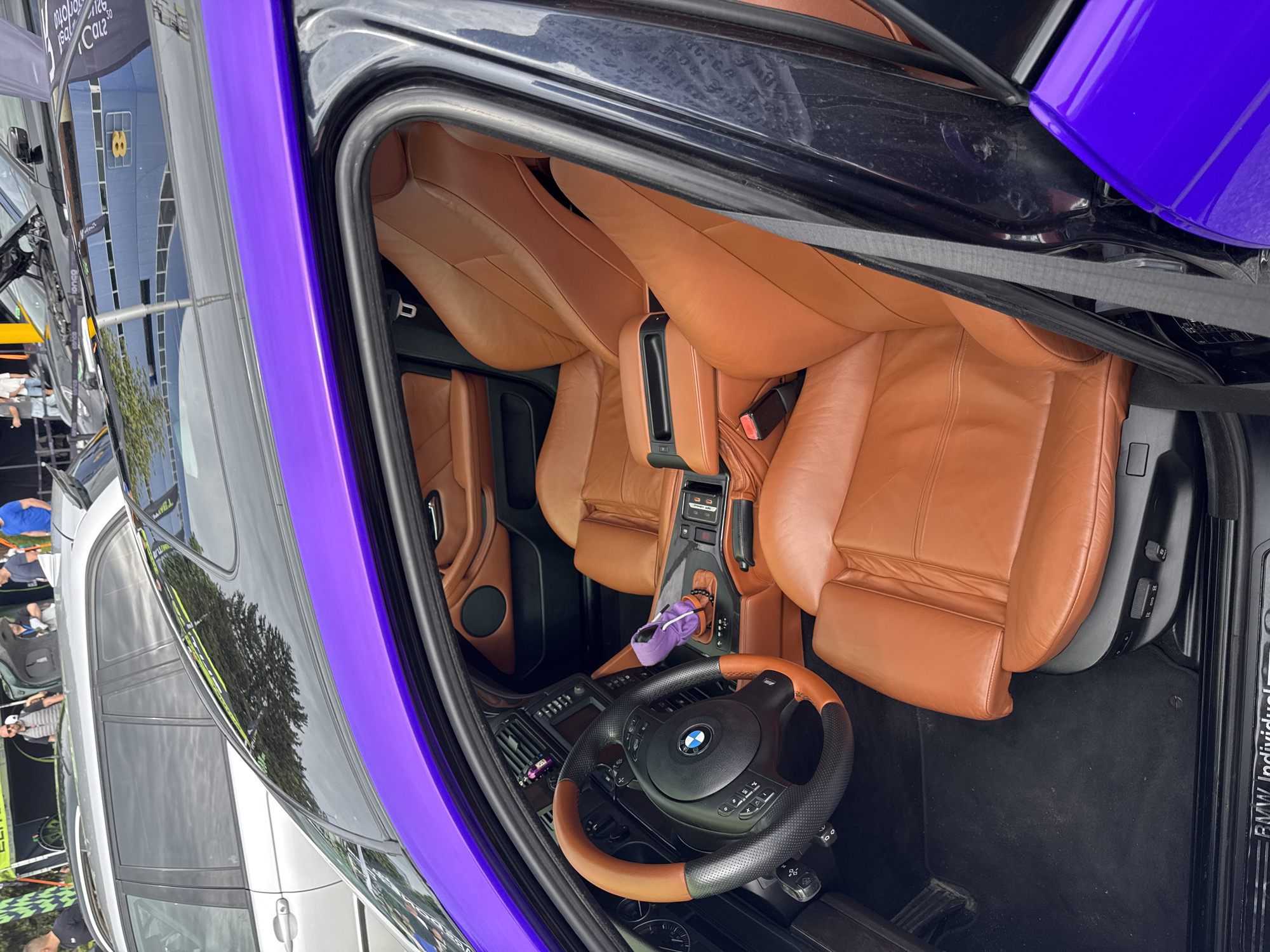 The image shows the interior of a BMW car, likely in Plovdiv, Bulgaria. The car features brown leather seats, a black dashboard, and a steering wheel with a brown accent. The car door is open, revealing a purple exterior accent on the door frame. A purple pouch sits in the center console area. The background shows reflections of buildings and trees in the car's windows, along with the presence of other cars and people in the distance, suggesting the car is parked in a public area, such as a car show or event. The environment appears to be outdoors on a sunny day.