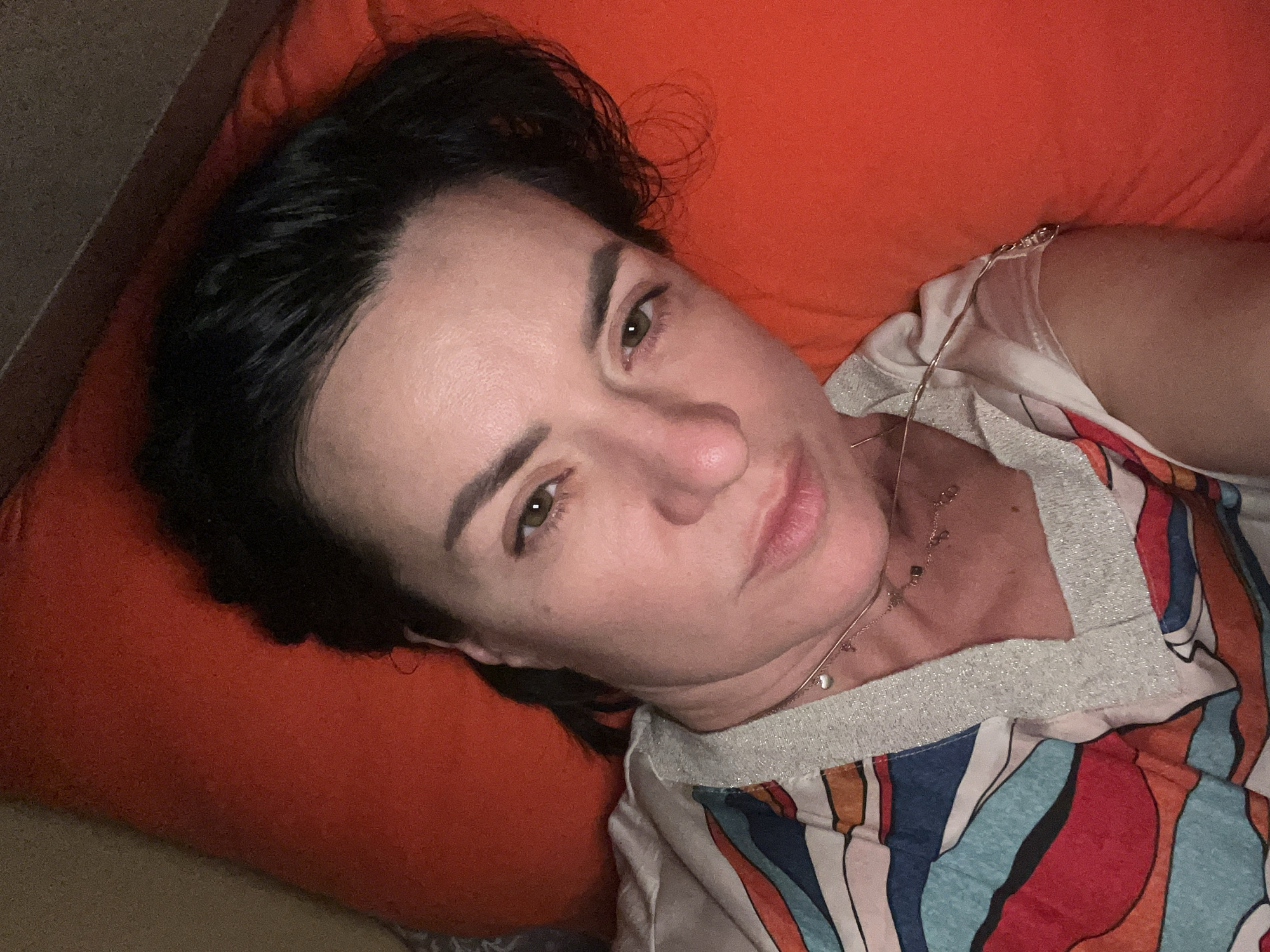 A woman with dark hair lies on an orange pillow, angled slightly towards the camera. She wears a patterned shirt with silver trim. A necklace with a small pendant is visible around her neck. The background suggests an interior setting. The location is not discernable from the provided image.