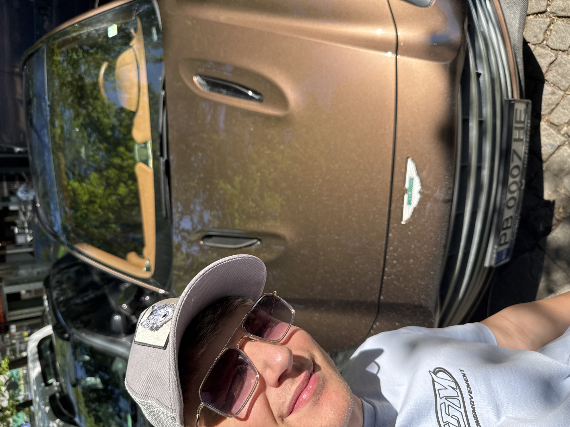 A close-up, medium-shot photograph shows a person posing in front of a brown Aston Martin car with a license plate that reads "PB 0007 HE." The person is wearing a gray baseball cap with a patch, and sunglasses with a golden frame. They are wearing a light blue t-shirt with black text. The car's front grill, headlights, and hood are clearly visible. The location is suggested to be the city of Plovdiv, Bulgaria, although this cannot be confirmed solely from the image. The photo is taken at an angle, with the car positioned vertically in the frame. The foreground is occupied by the person. The background includes additional cars.