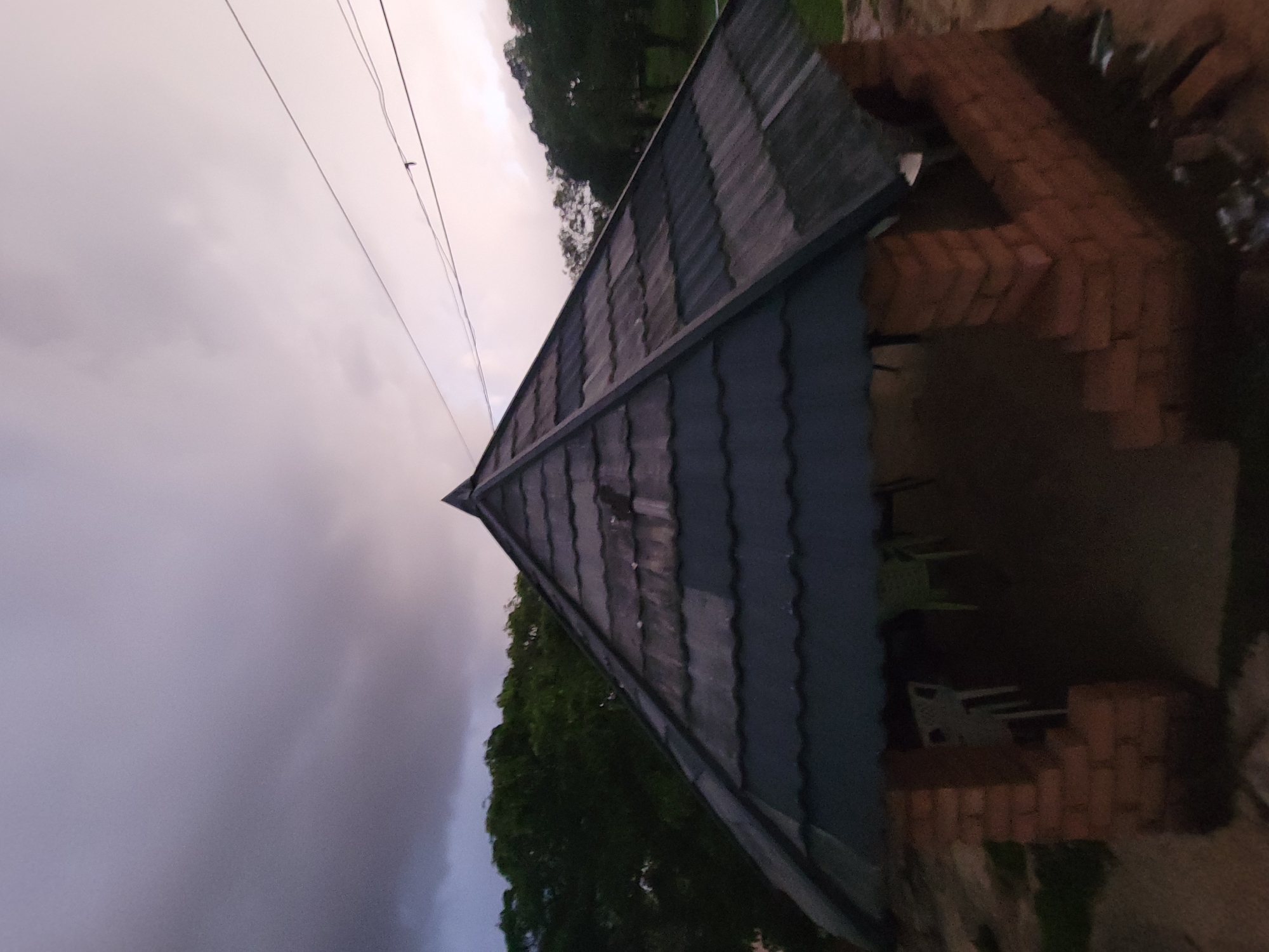The image depicts a pyramidal-roofed shelter or gazebo under an overcast sky. The structure features a dark grey or blue corrugated metal roof, visibly segmented into panels that converge at a central peak. Its base is constructed from reddish-brown brick pillars, forming low walls with open sections. Inside the dimly lit interior, at least two chairs are discernible: one light-colored, possibly white, and another green.
In the background, dense green foliage from trees is visible behind the left side of the shelter. The sky exhibits varying cloud density and color; the upper left quadrant is dominated by darker, heavy grey clouds, while the upper right transitions to lighter, pale grey and faint pink tones. Multiple thin, dark power lines traverse the upper right portion of the sky, with one line featuring a small, dark connector or insulator. The ground surrounding the structure appears to be uneven dirt or earth, possibly with sparse vegetation. No individuals or animals are present in the scene. The overall lighting suggests twilight, dusk, or heavily overcast conditions. The exact location is unknown.