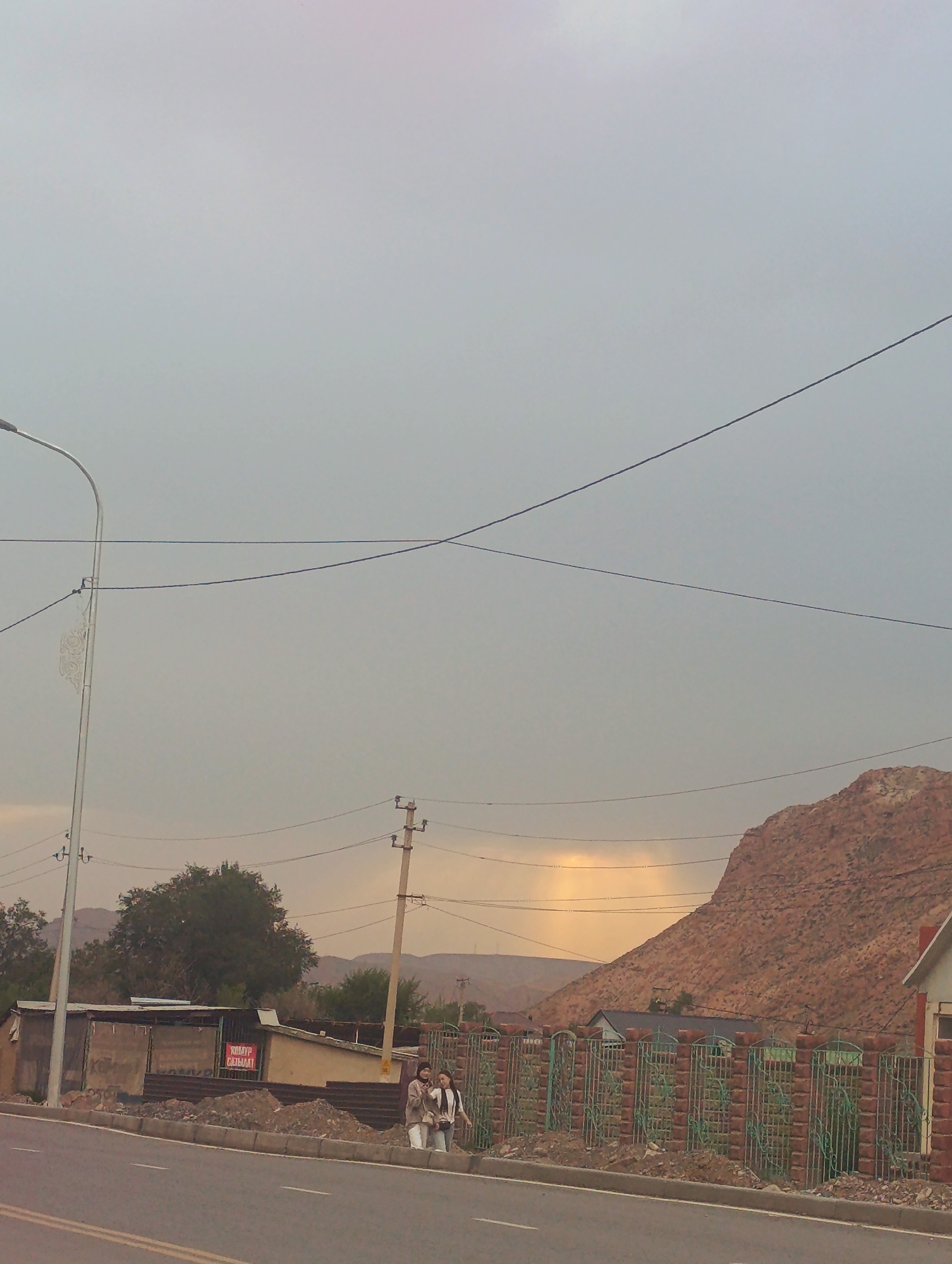The image captures a street scene in Naryn City, Kyrgyzstan. In the foreground, a paved road with white lane markings is visible. To the right of the road, a green metal fence with brick pillars encloses an area. Beyond the fence, a large, reddish-brown mountain dominates the background. The sky is overcast with a hint of sunlight breaking through in the distance, creating a yellowish hue.
Along the roadside, there are two women walking on the sidewalk. One woman is wearing a light-colored hijab and a beige jacket, while the other is wearing a white hijab and a dark jacket. They are walking away from the viewer, towards the right side of the image.
Behind the women, a low-rise building with a corrugated metal roof is situated. A red sign with Cyrillic lettering is attached to the building. A lamppost with decorative elements stands to the left of the building. A utility pole with wires is positioned between the women and the building. Trees are sparsely scattered in the middle ground, and a distant mountain range is visible on the horizon. Power lines crisscross the sky, and a street lamp is present on the left side of the image.