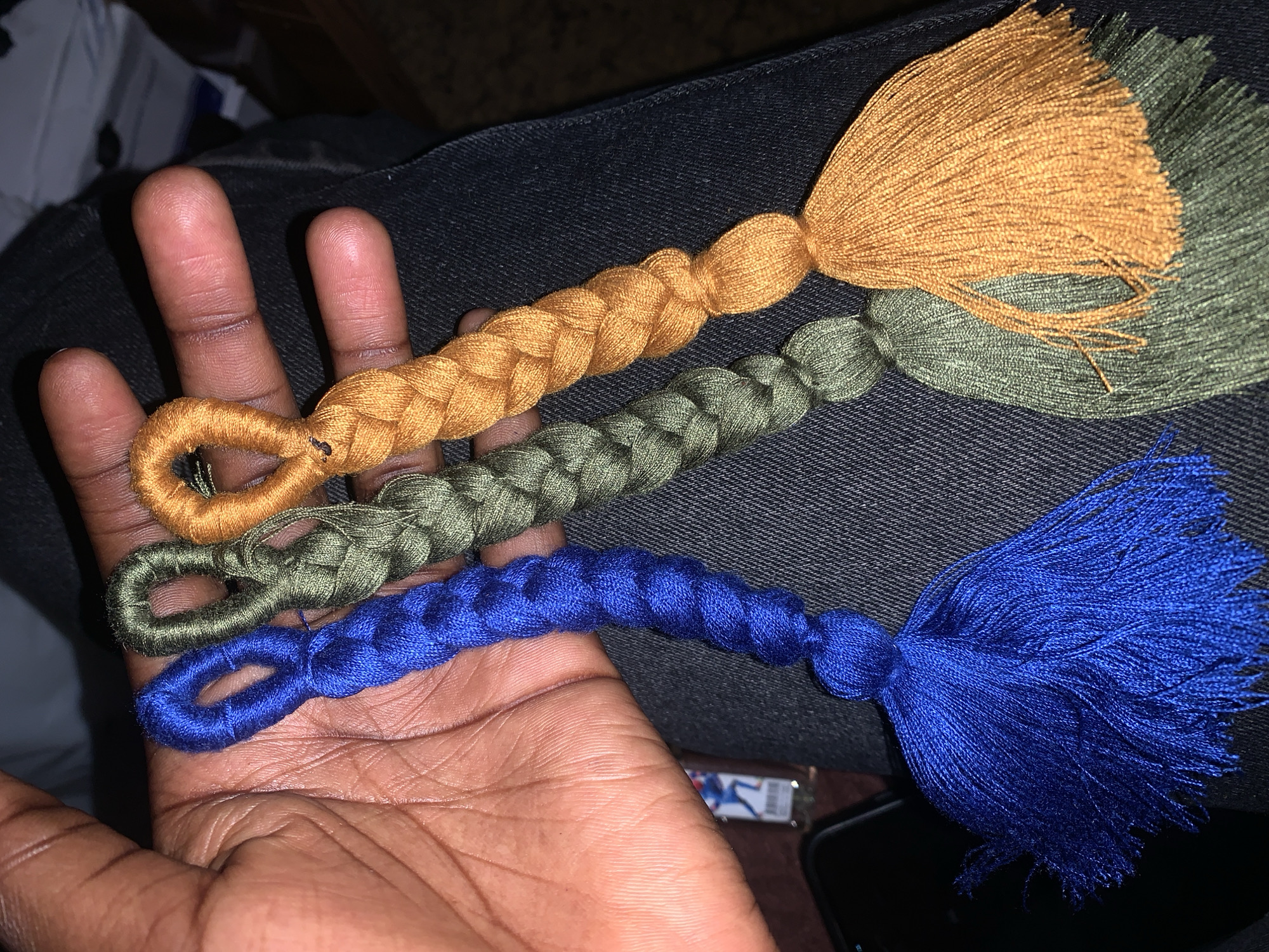 A close-up, overhead shot shows a person's hand holding three braided tassels in various colors: royal blue, olive green, and orange. The tassels are made of thick, yarn-like material and have a loop at the top for hanging. They are held in a relaxed grip, with the fingers partially curled around them. The background consists of dark denim fabric, likely pants. A dark smartphone and a portion of a lighter are visible on the lower left of the image. The scene is dimly lit, creating a warm and intimate atmosphere. There is no identifiable location in the city of Gura Top, Nigeria.